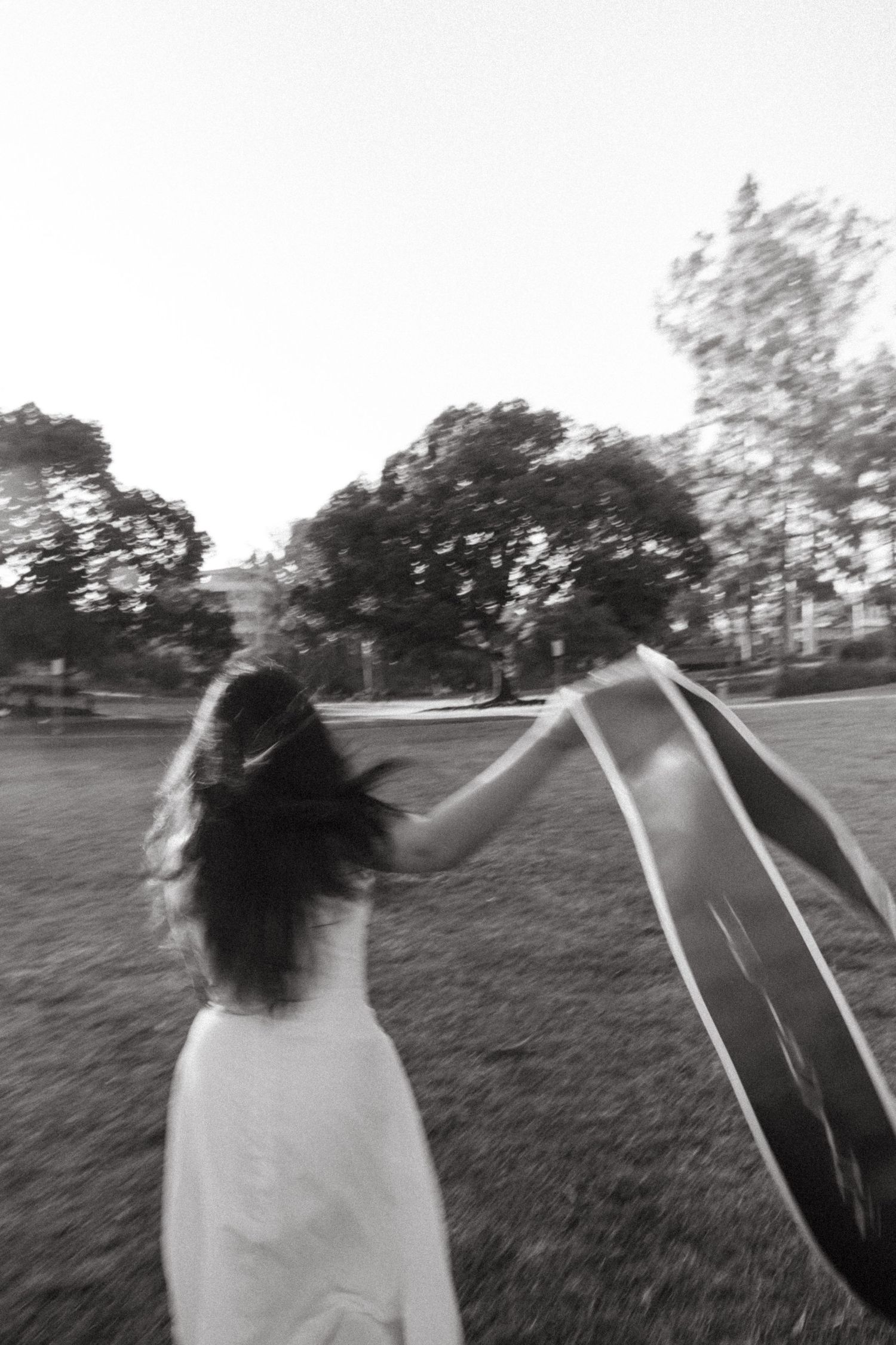 Black and white photo of woman in white dress spinning with graduation sash in outdoor park.