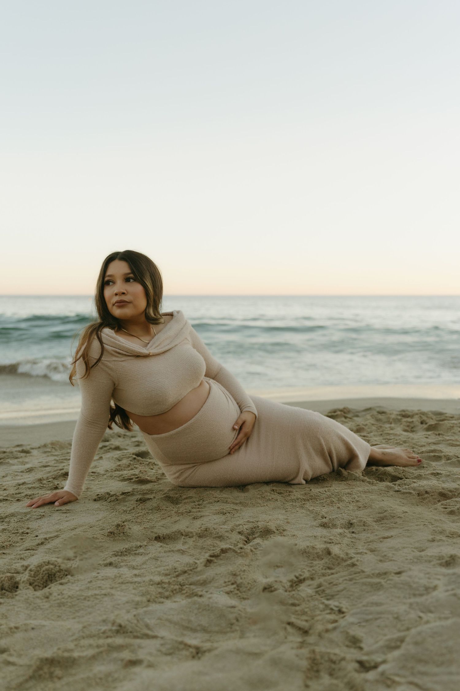 Pregnant woman in beige two-piece outfit poses on sandy beach at dusk with ocean behind her.
