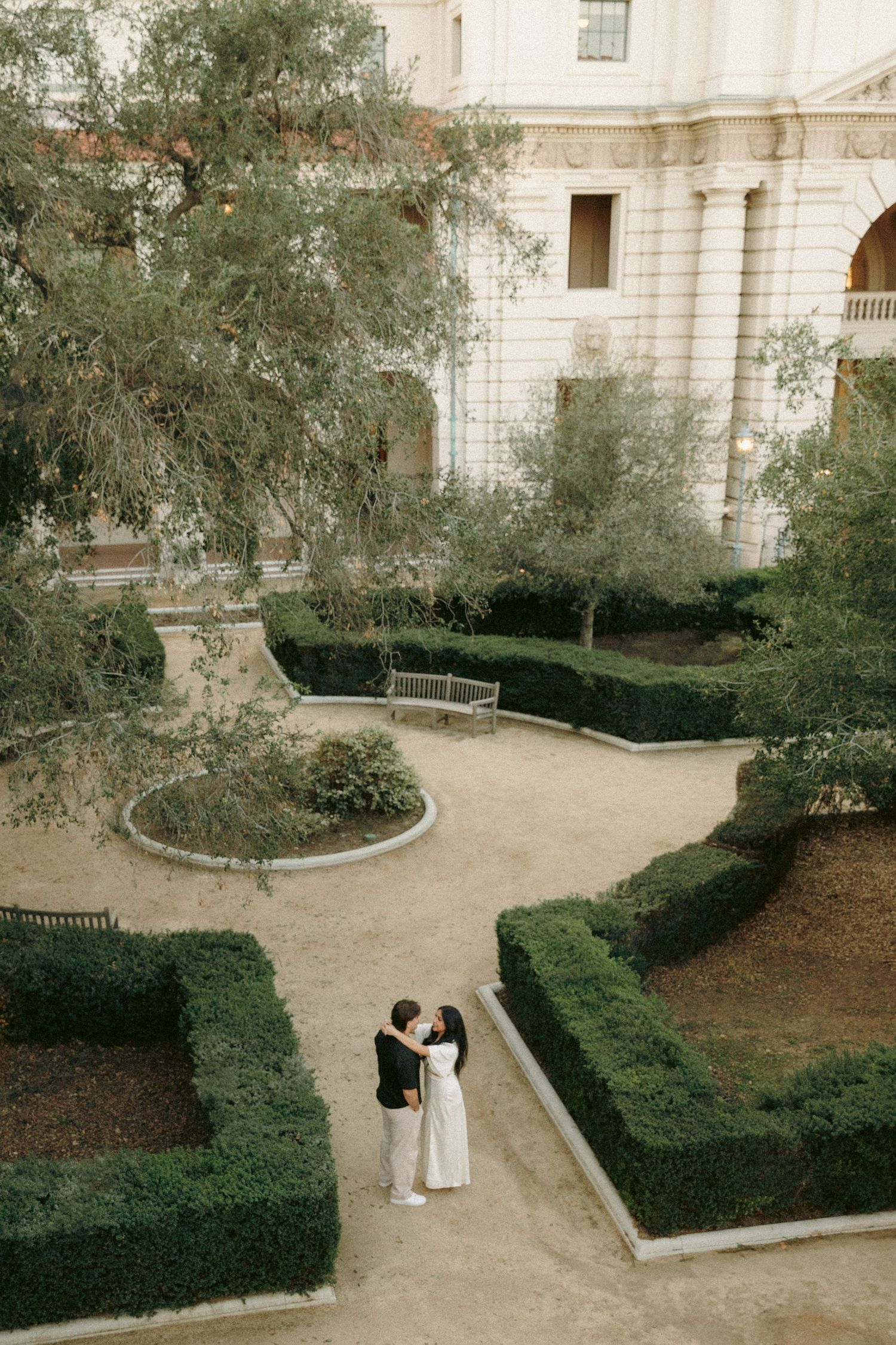 Aerial view of couple embracing in a formal garden courtyard with manicured hedges and elegant architecture.