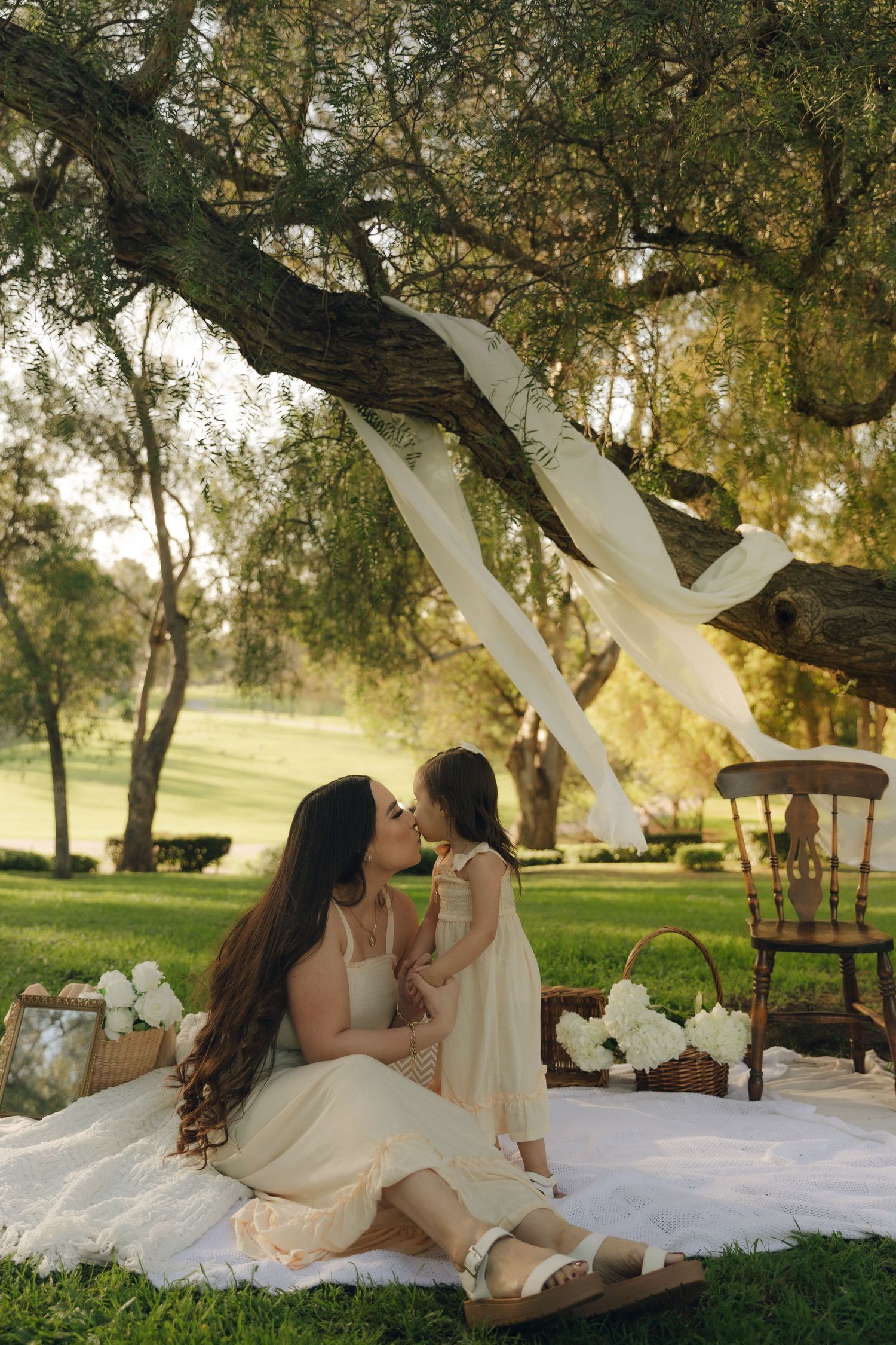 Mother kissing young daughter on picnic blanket under large tree in sunlit green park.