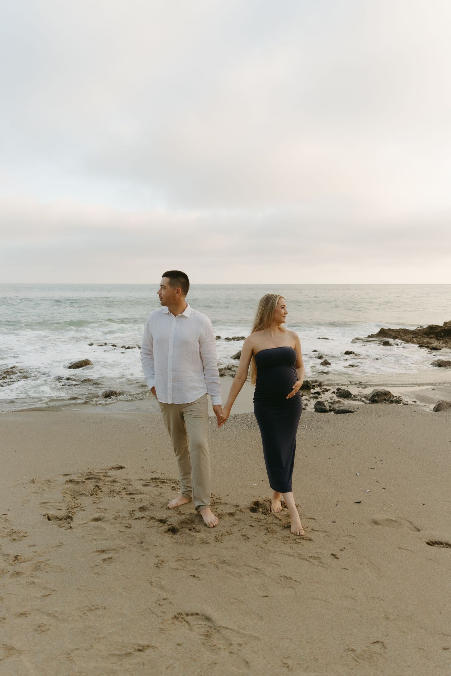 Pregnant couple holding hands walking along ocean beach at dusk, woman in black dress, man in white shirt.