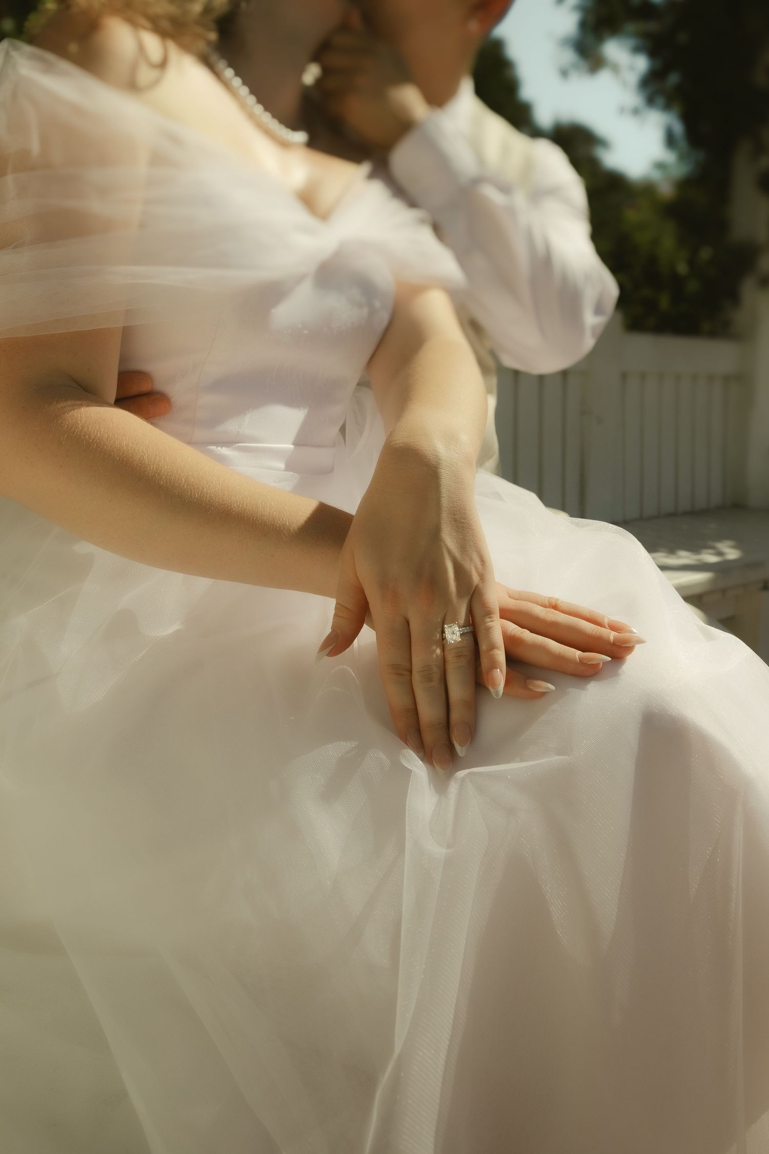 Close-up of a bride in a white tulle gown displaying her diamond engagement ring while seated outdoors.