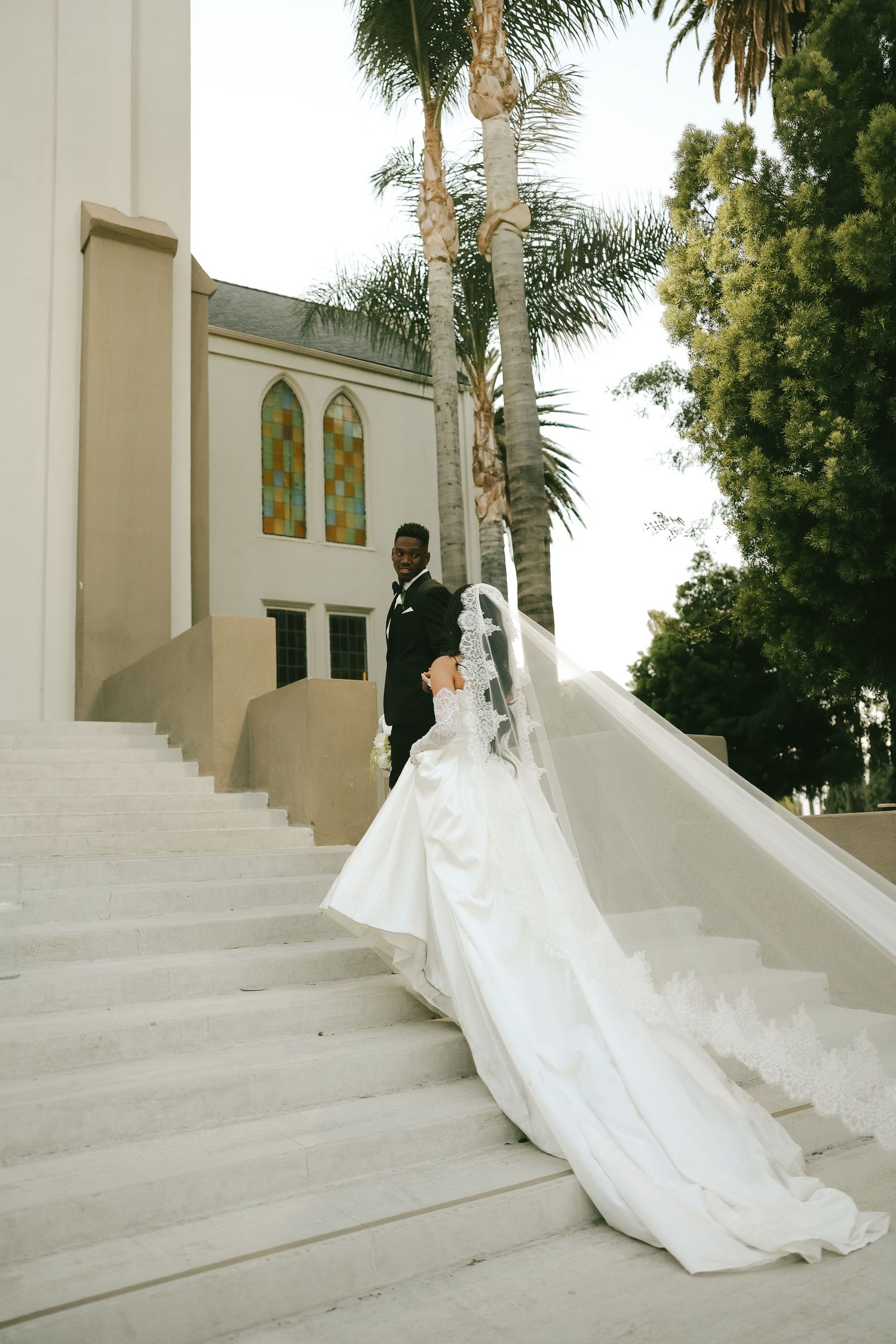 Bride in dramatic ballgown with long train descends grand white church steps as groom in black tuxedo watches.