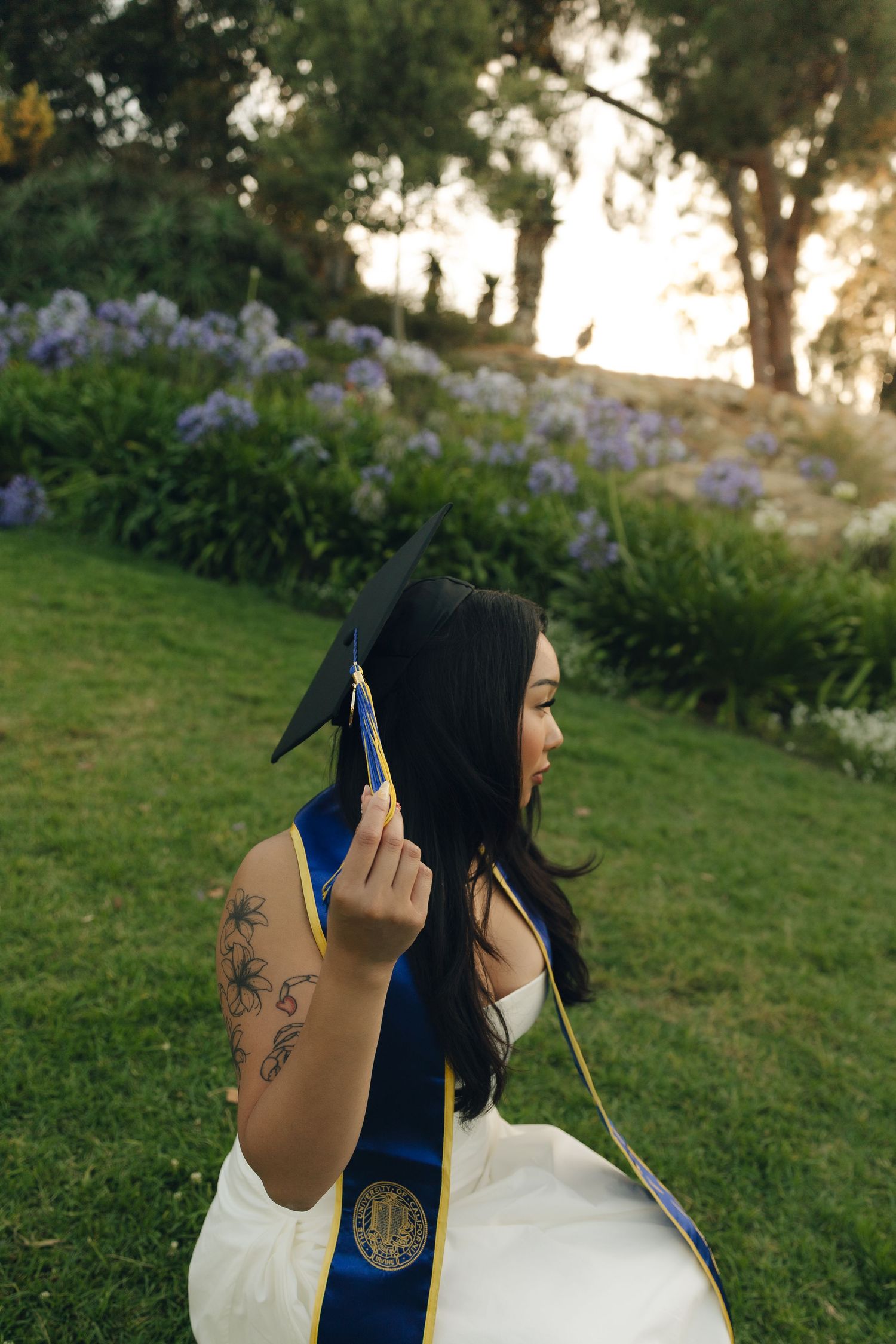 Graduate in white dress holding mortarboard cap with blue sash seated near purple flowers at sunset.