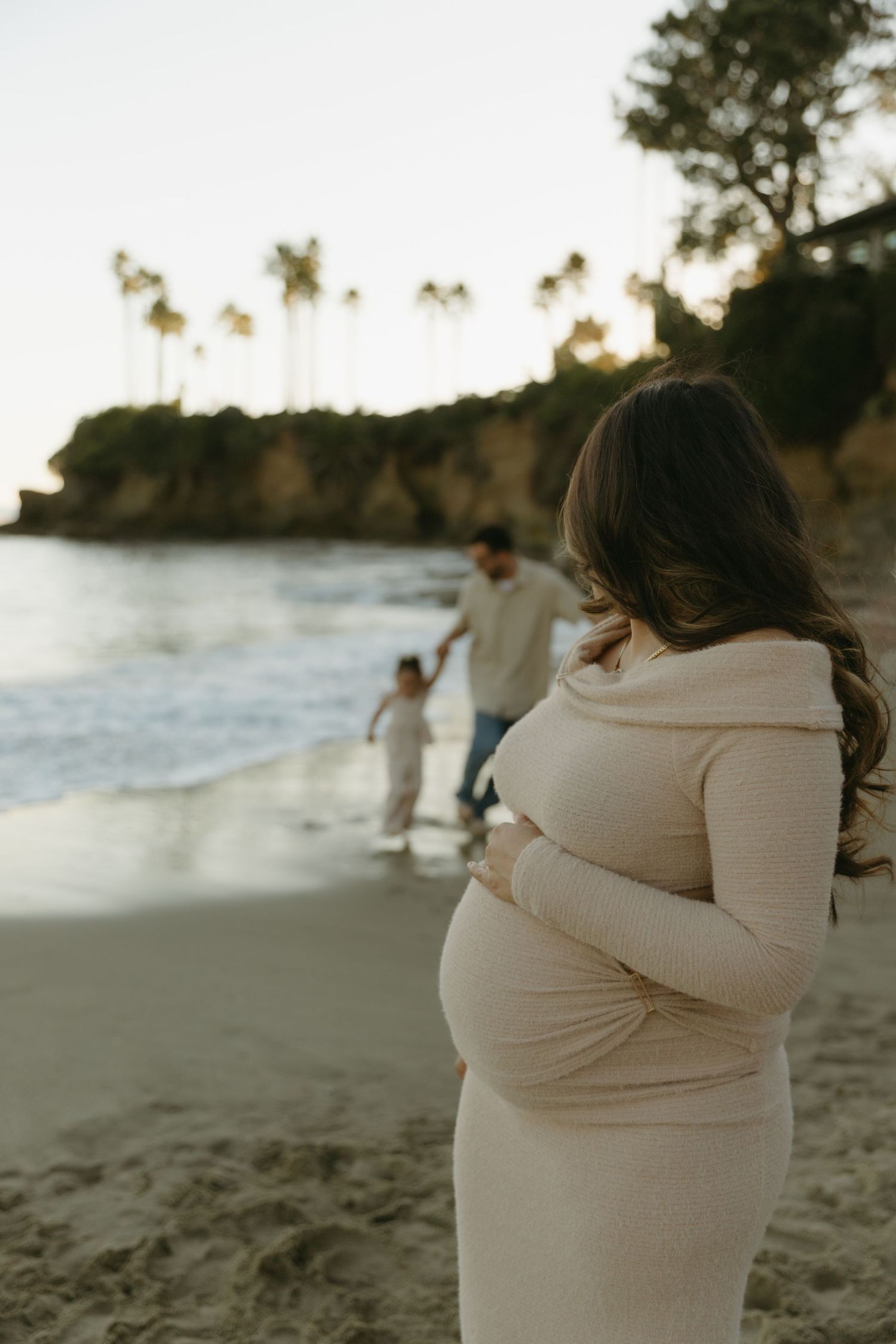 Pregnant woman in beige dress holds belly on beach while family plays near water at sunset.
