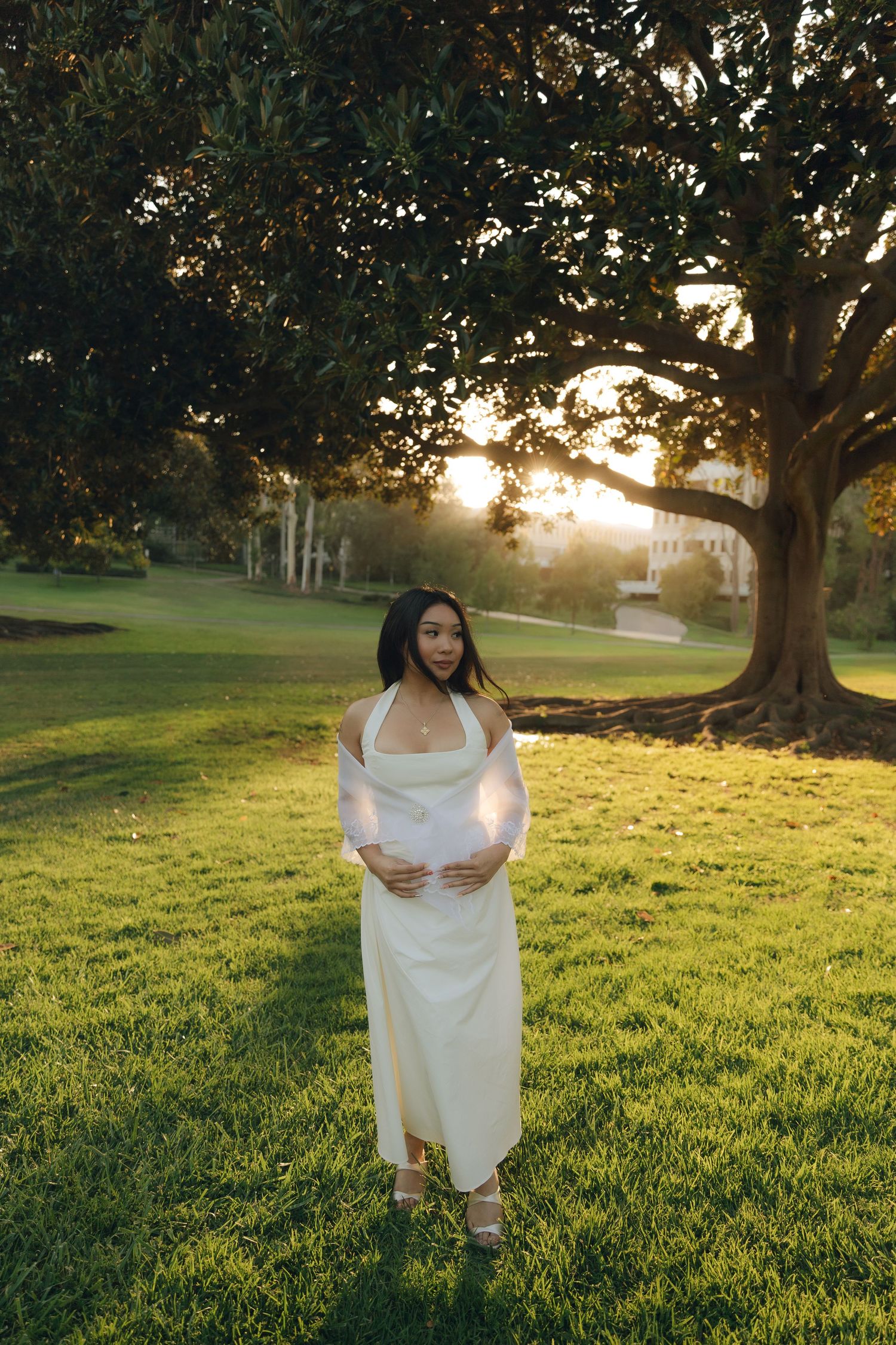 Pregnant woman in white dress holds belly while standing on green lawn under large tree at sunset.