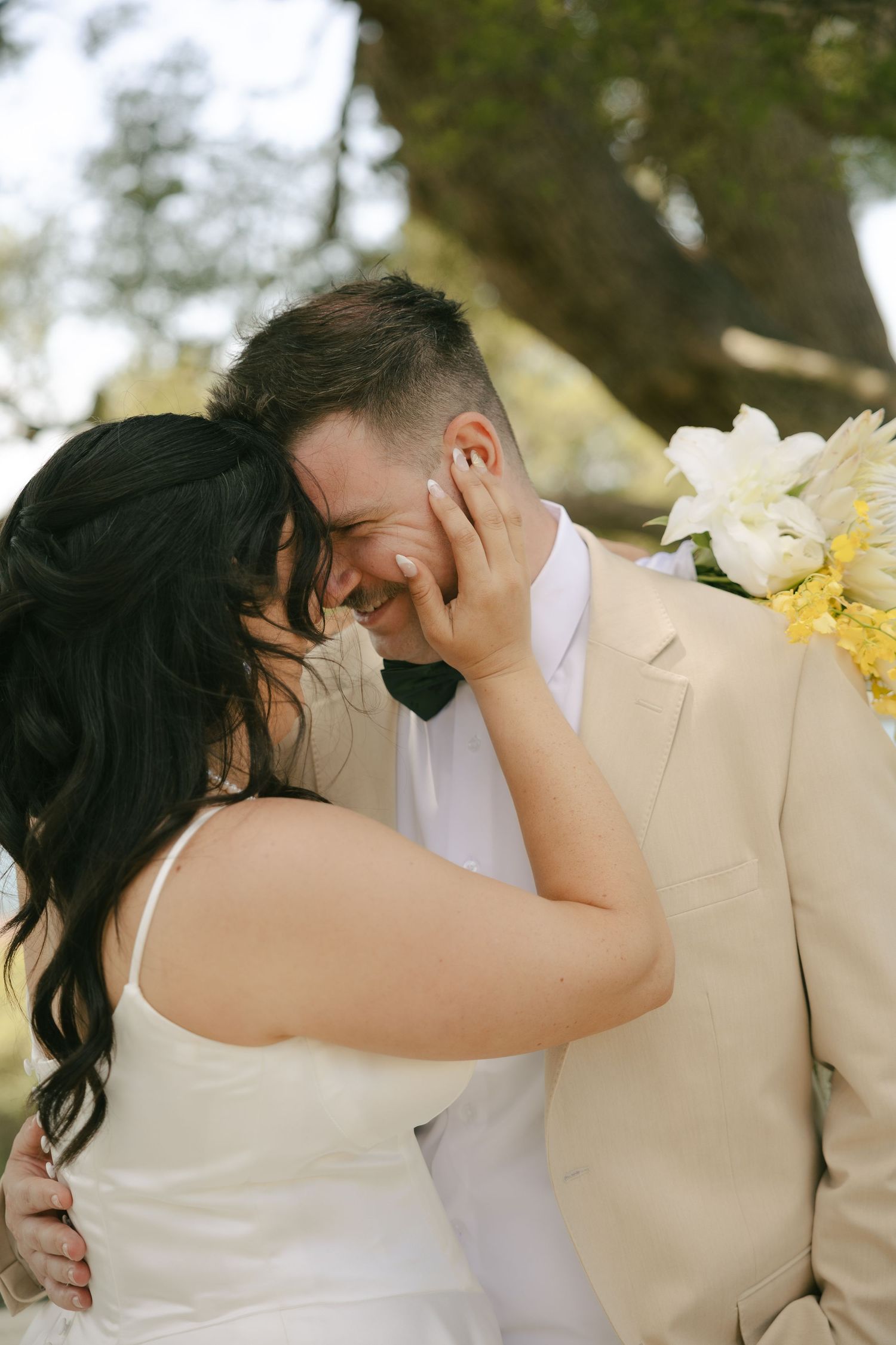 Emotional bride and groom sharing tender moment laughing together outdoors under trees.