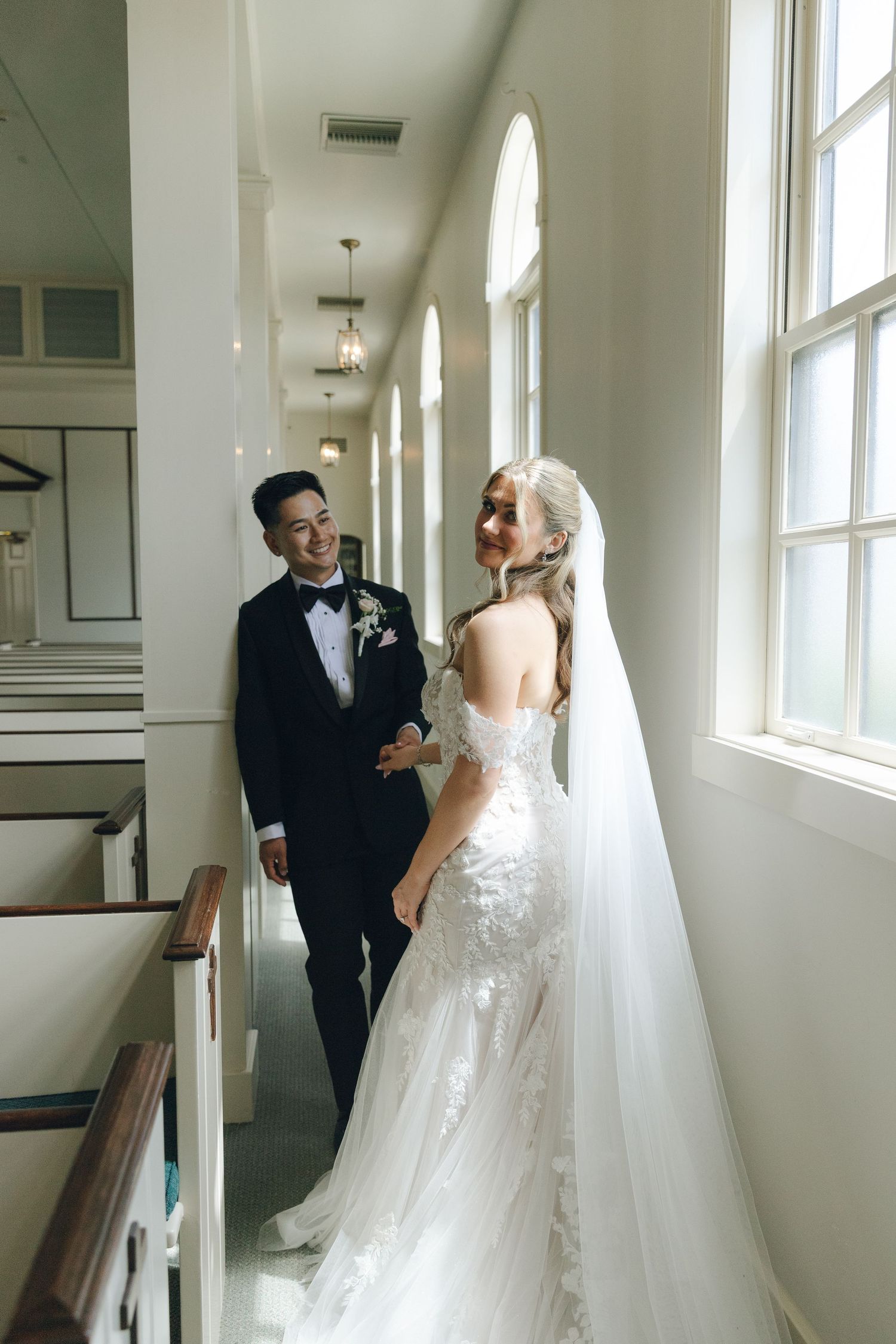 Smiling groom in black tuxedo gazes at bride in lace mermaid gown inside a sunlit white church.