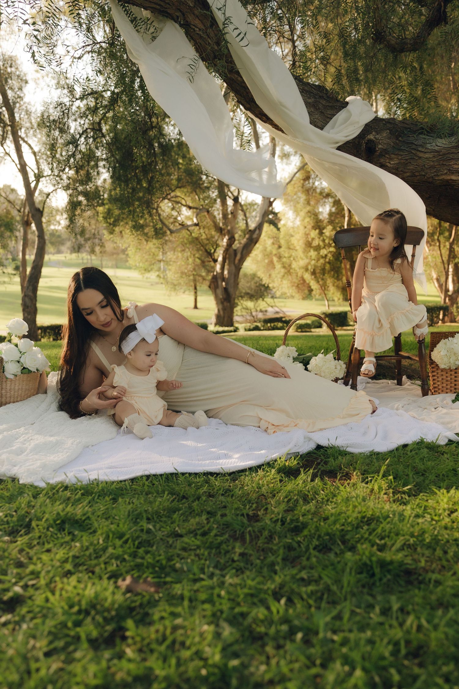 Mother in cream dress relaxing on blanket with baby while toddler stands nearby under tree.