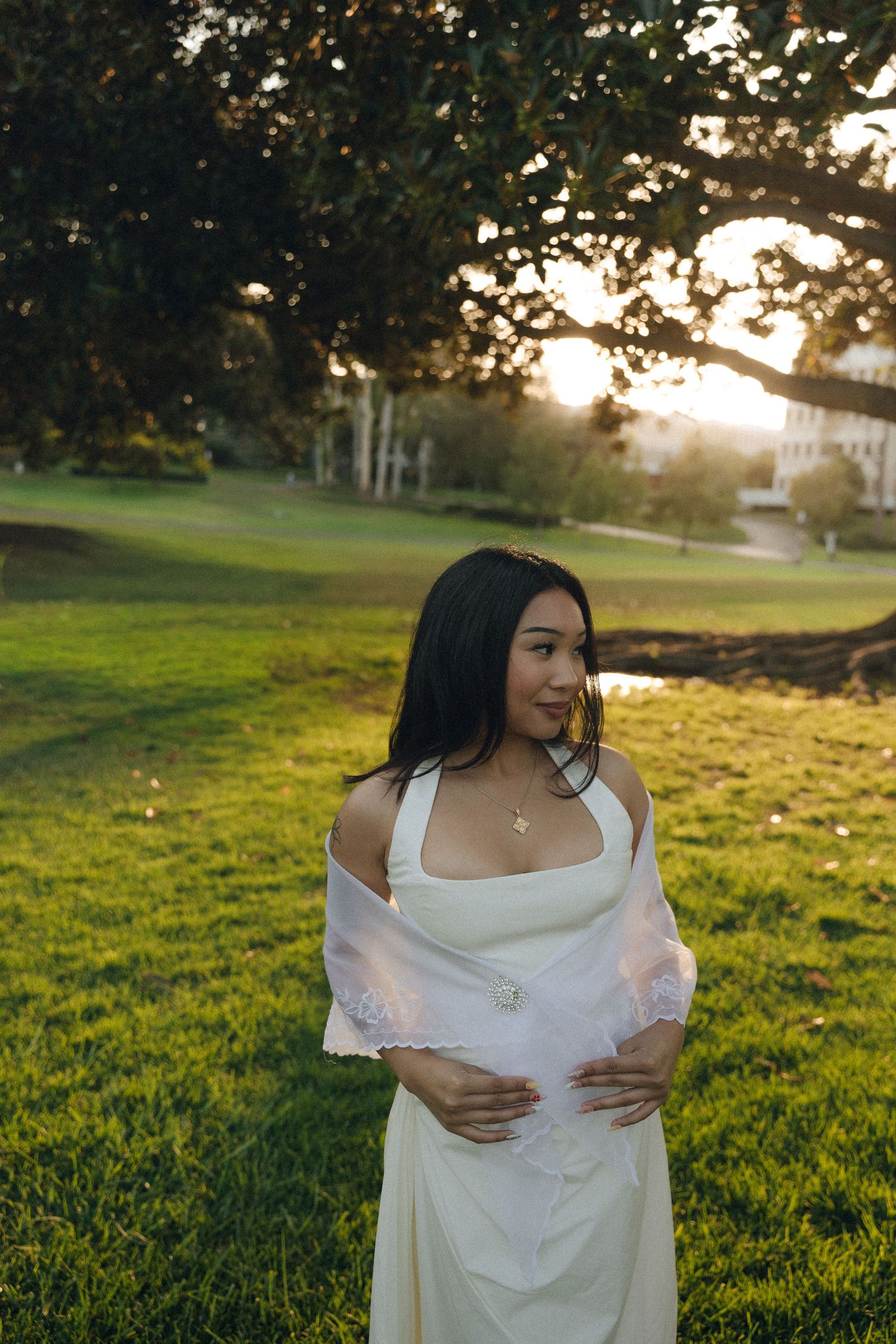Young woman in white off-shoulder dress standing on green lawn during warm golden sunset light.