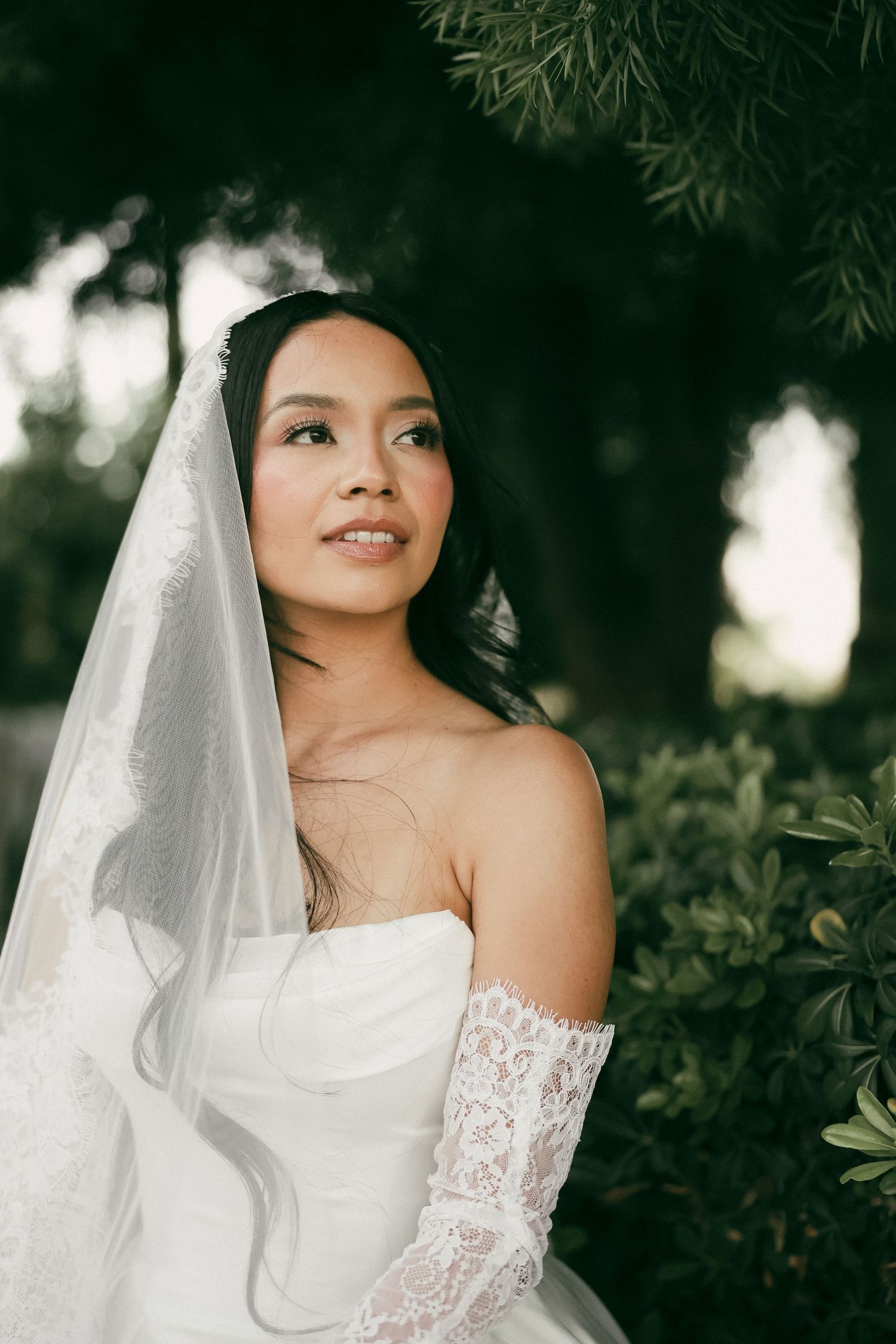 Elegant Asian bride in strapless lace wedding gown and veil gazes upward surrounded by lush green foliage.