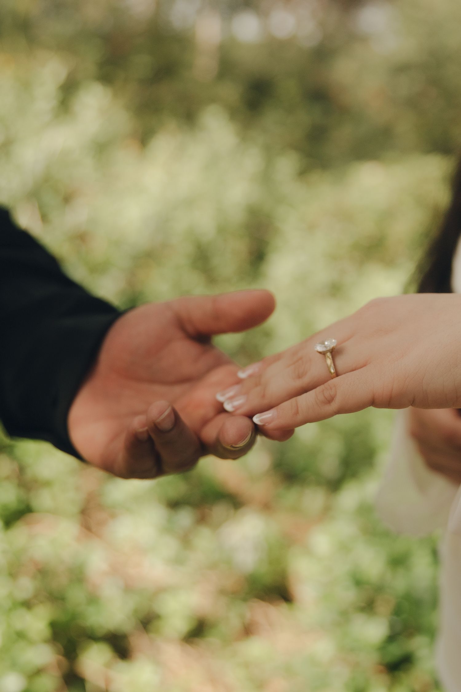 Close-up of couple holding hands showing elegant diamond engagement ring with green bokeh background.