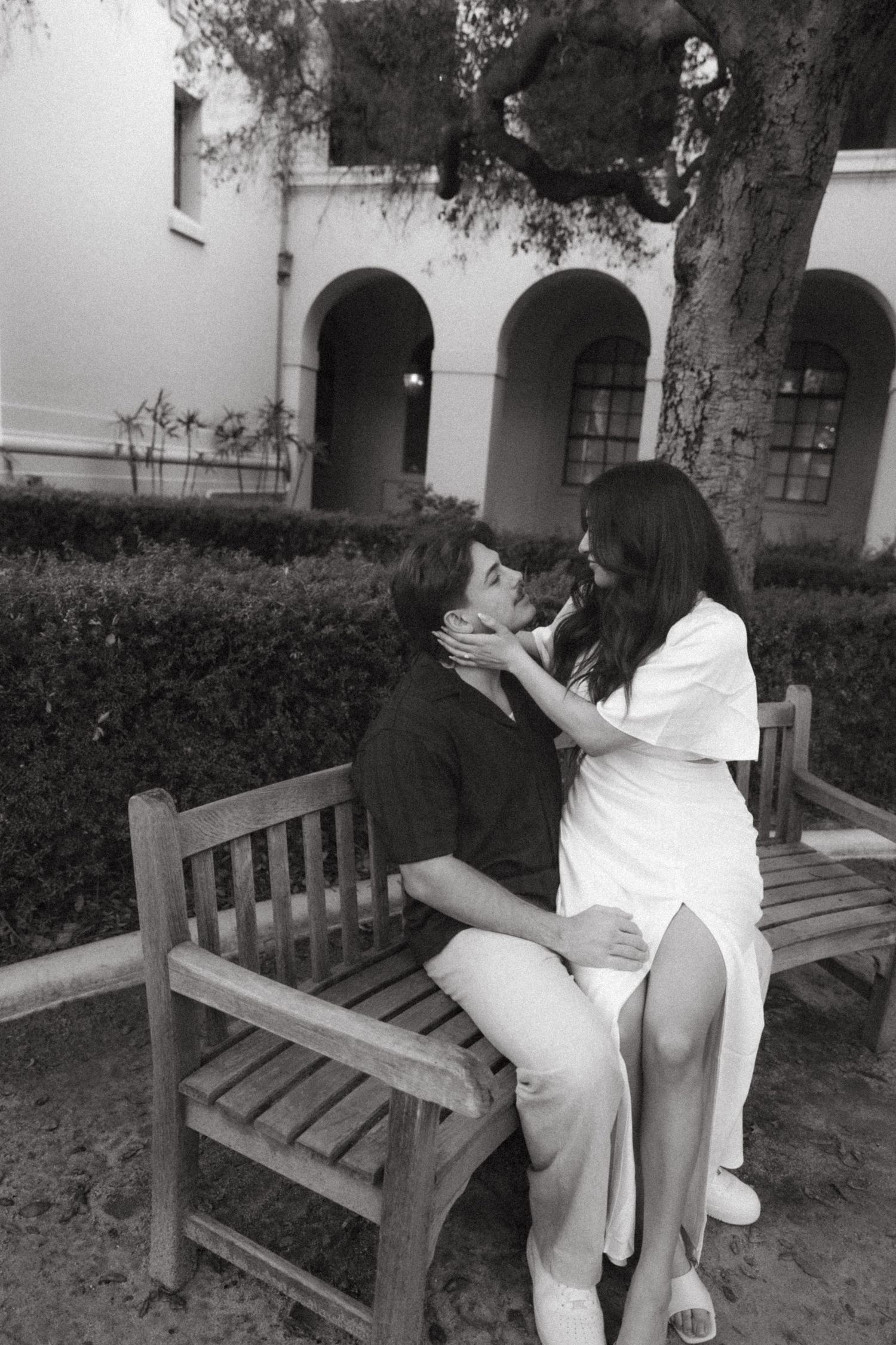 Black and white photo of a couple sharing a kiss on a wooden bench in a garden courtyard.