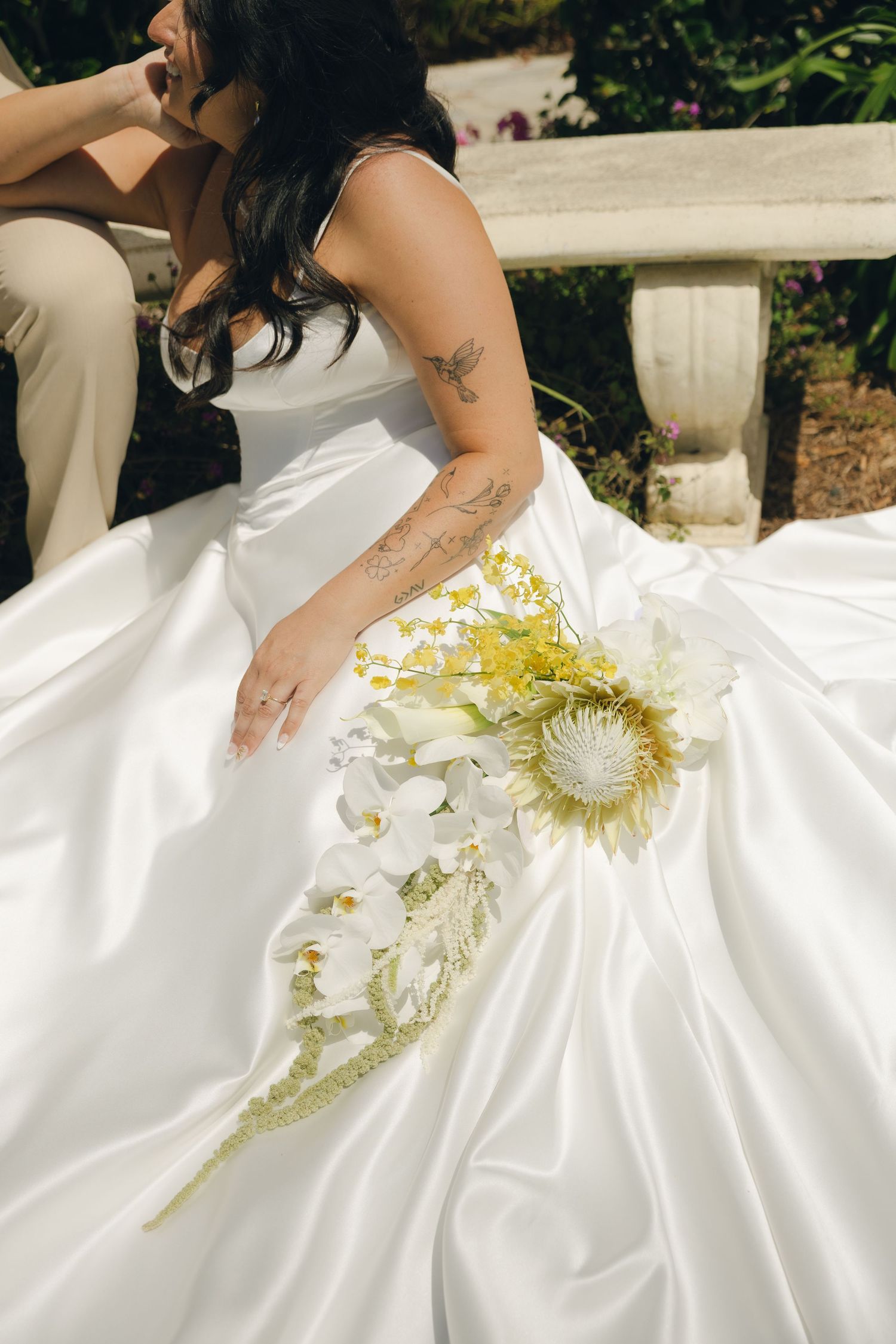 Bride in white satin gown holding white orchid and protea bouquet, tattoos visible on arm, seated outdoors.