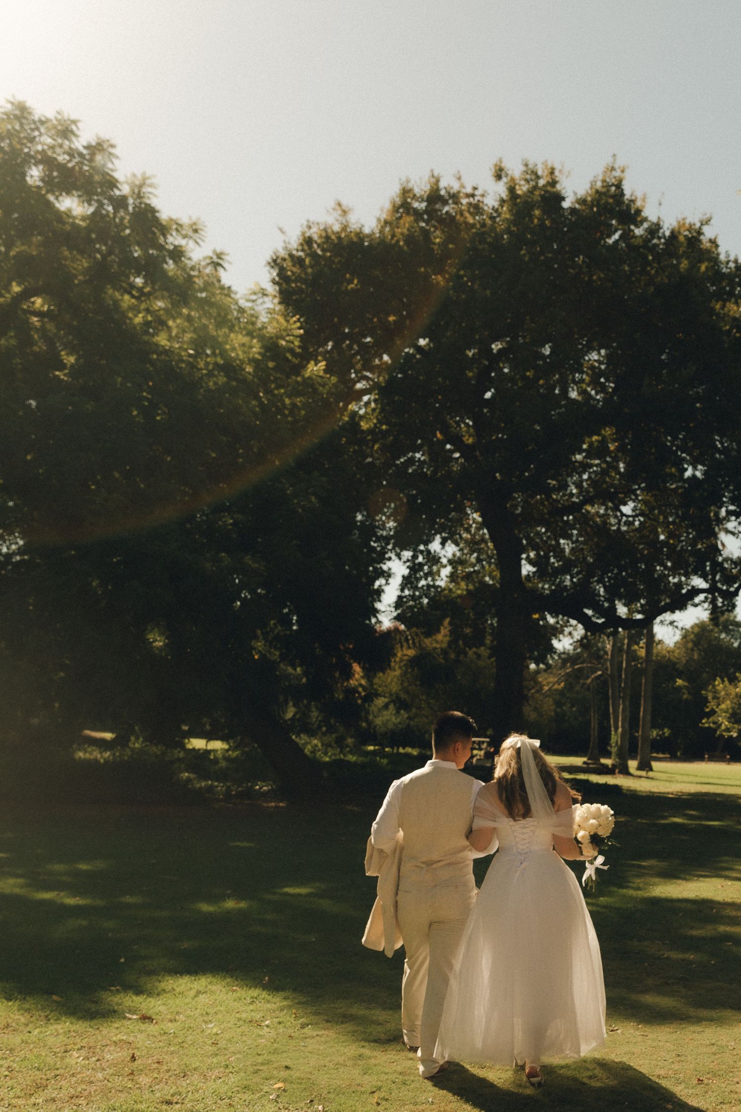 Bride and groom walking hand-in-hand through a sunlit park with lush green trees on their wedding day.