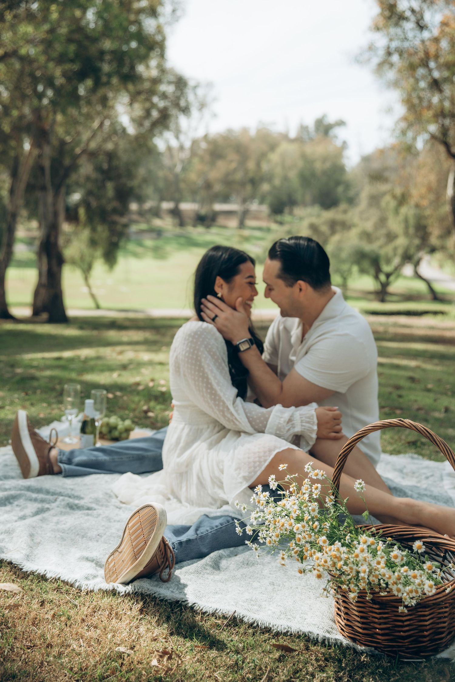 Loving couple almost kissing on a picnic blanket in a sunny park surrounded by trees and daisy flowers.