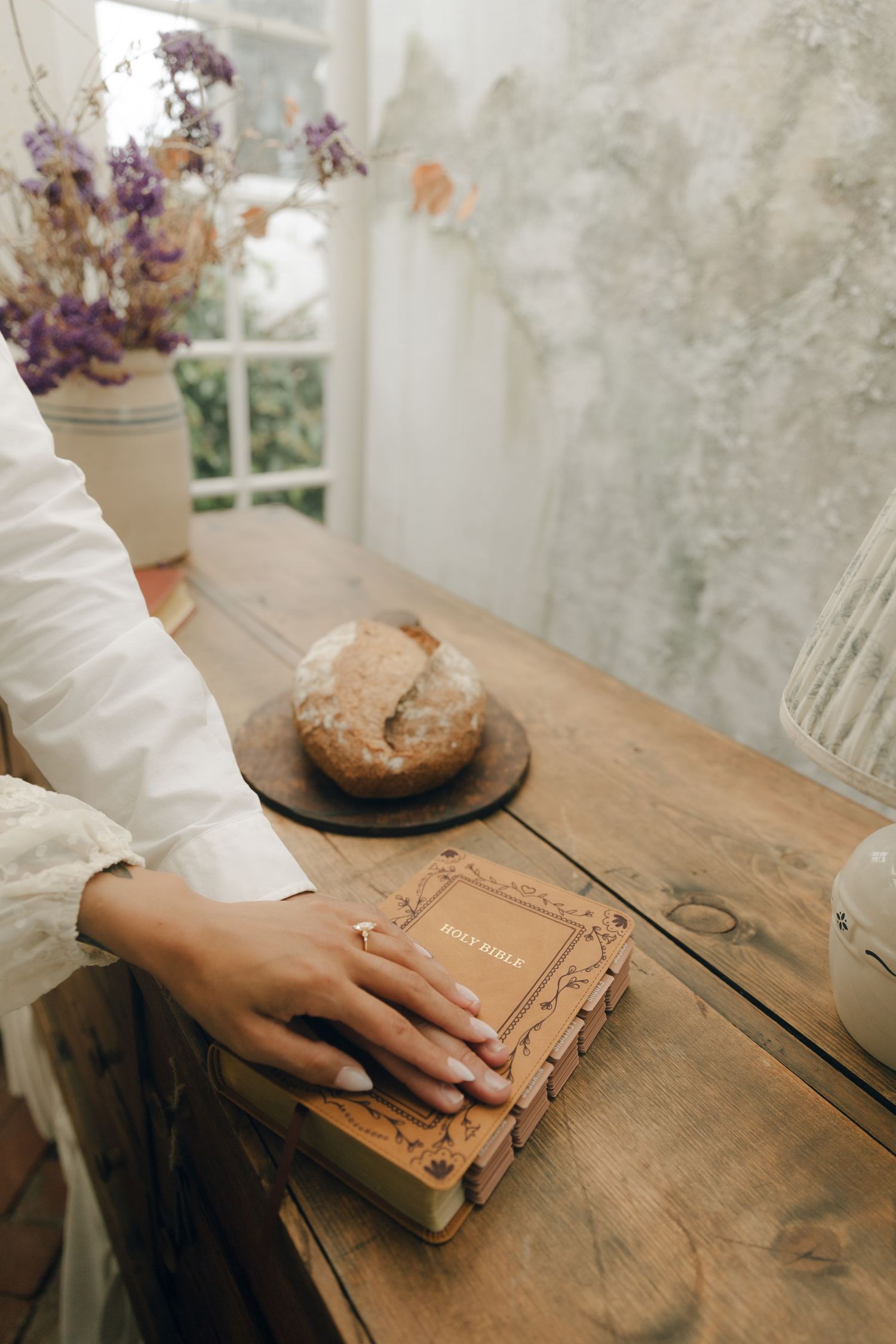 A hand rests on a Holy Bible on a rustic wooden table with bread and dried flowers nearby.