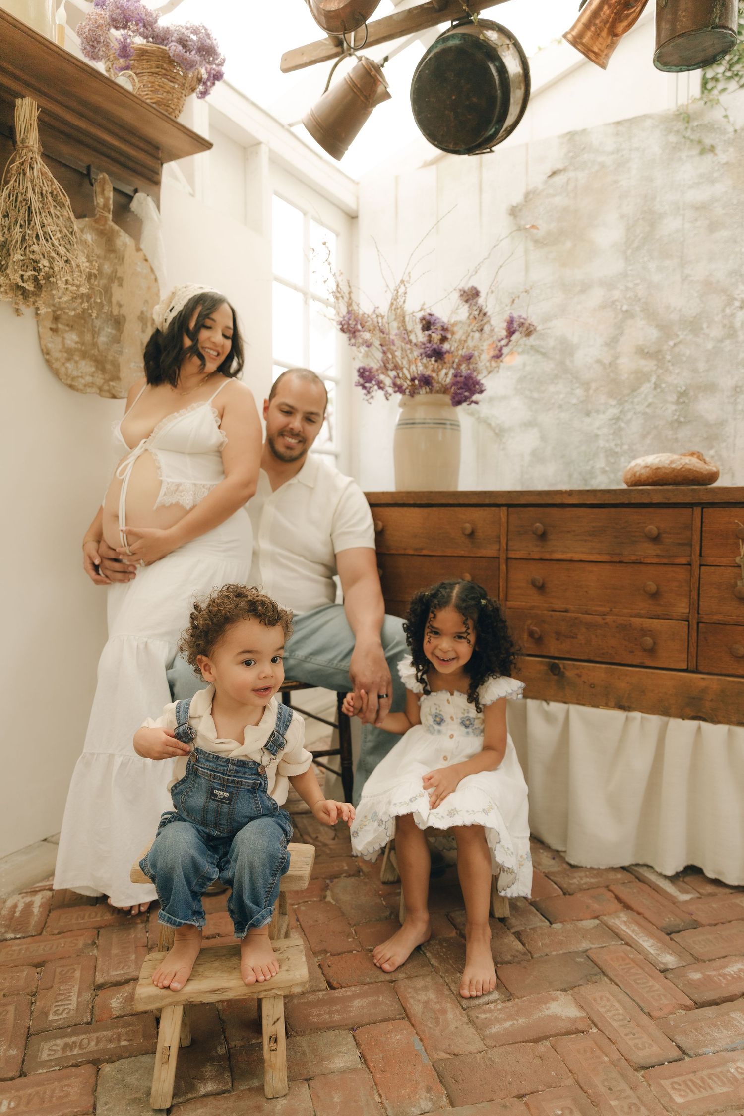 Pregnant mother and father pose with two young children in a rustic boho-styled indoor setting.