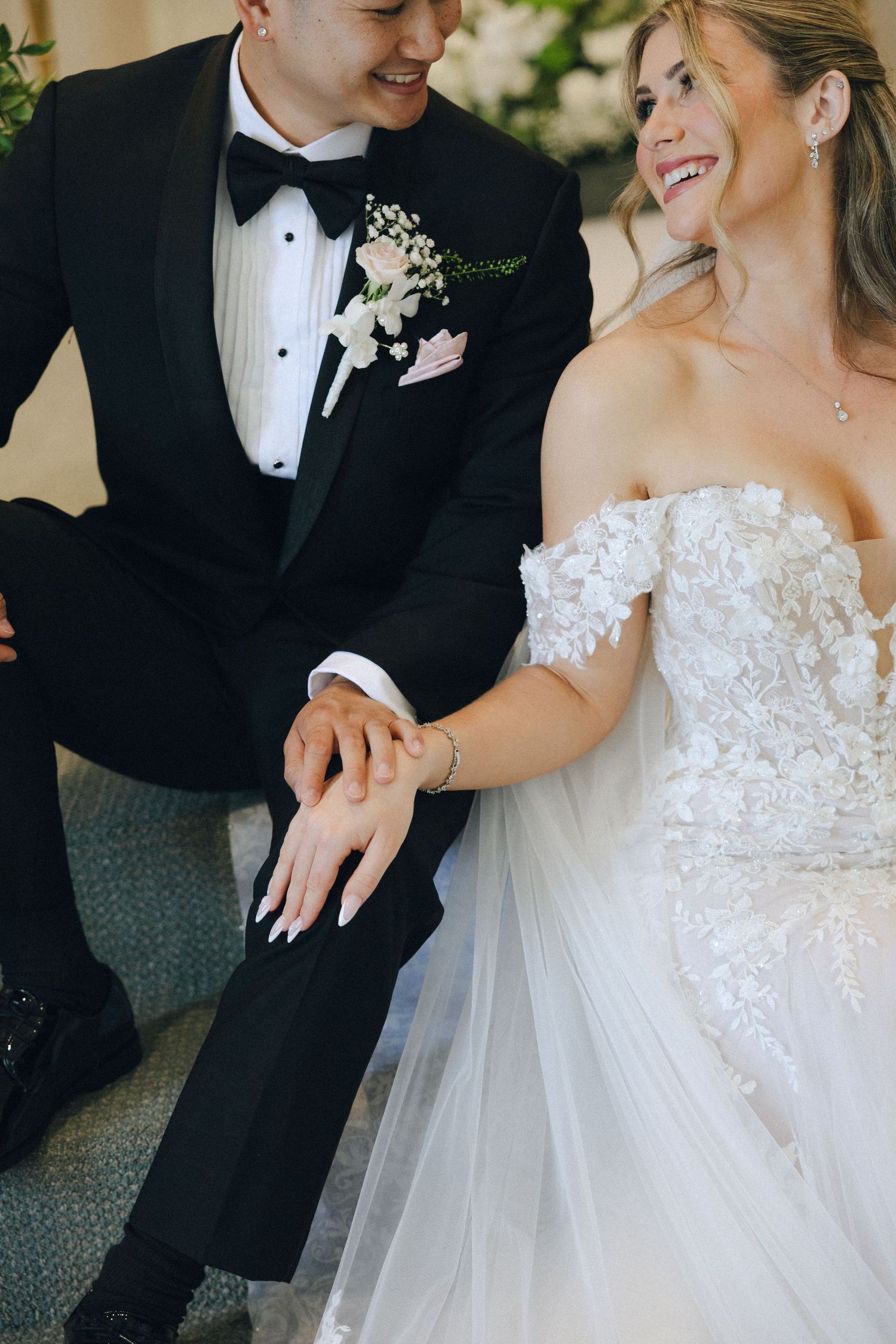 Laughing bride in off-shoulder lace gown sits beside groom in black tuxedo with floral boutonniere.