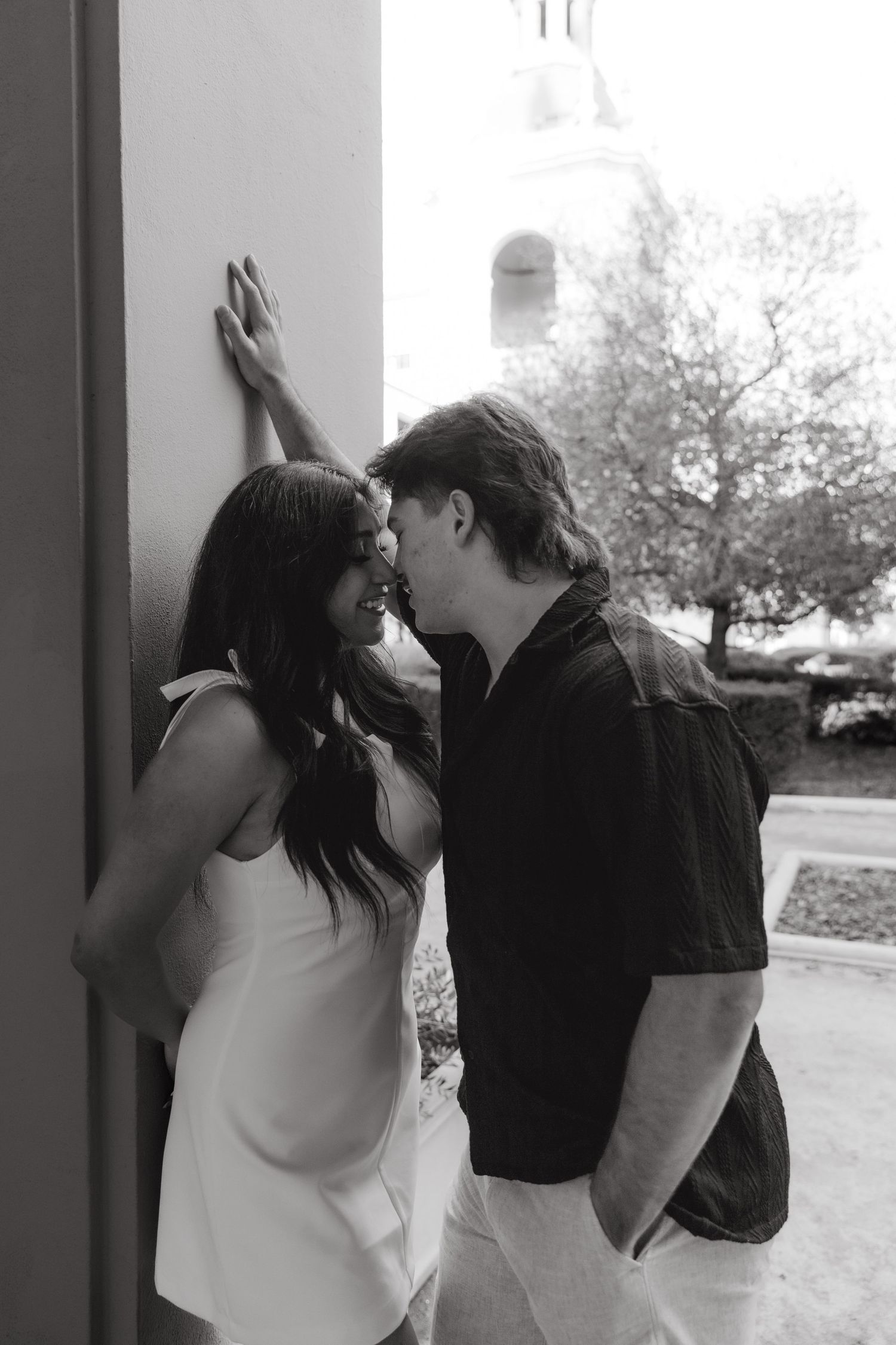 Black and white photo of a romantic couple leaning against a wall, almost kissing outdoors.