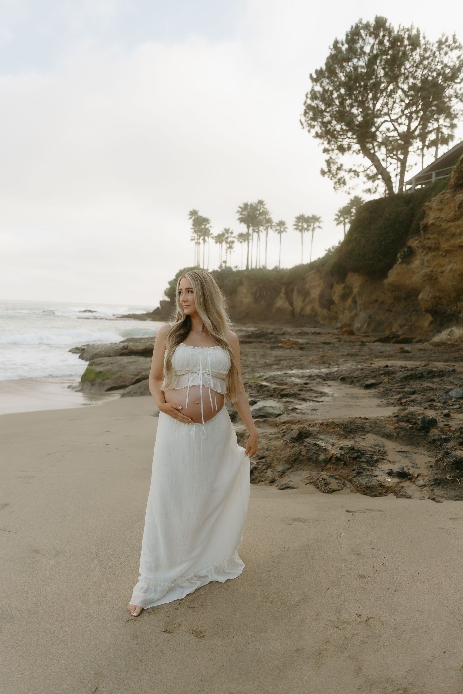 Pregnant blonde woman in white strapless gown standing on rocky beach with palm trees and cliffs behind her.