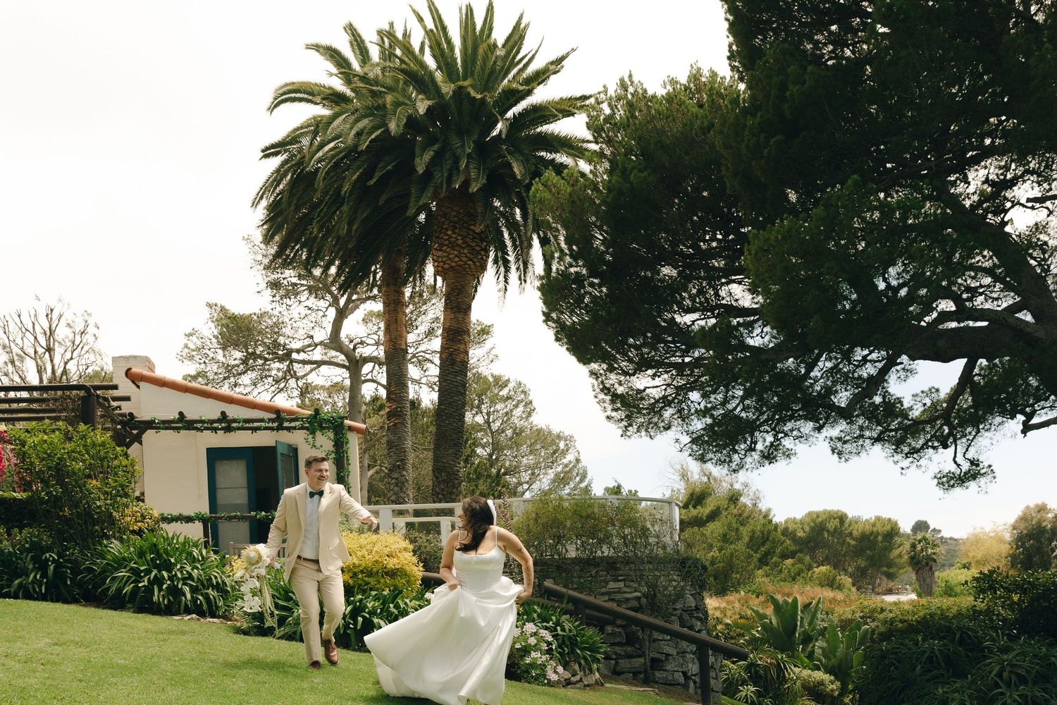 Bride and groom walking on lush green lawn with palm trees and garden backdrop at outdoor wedding venue.
