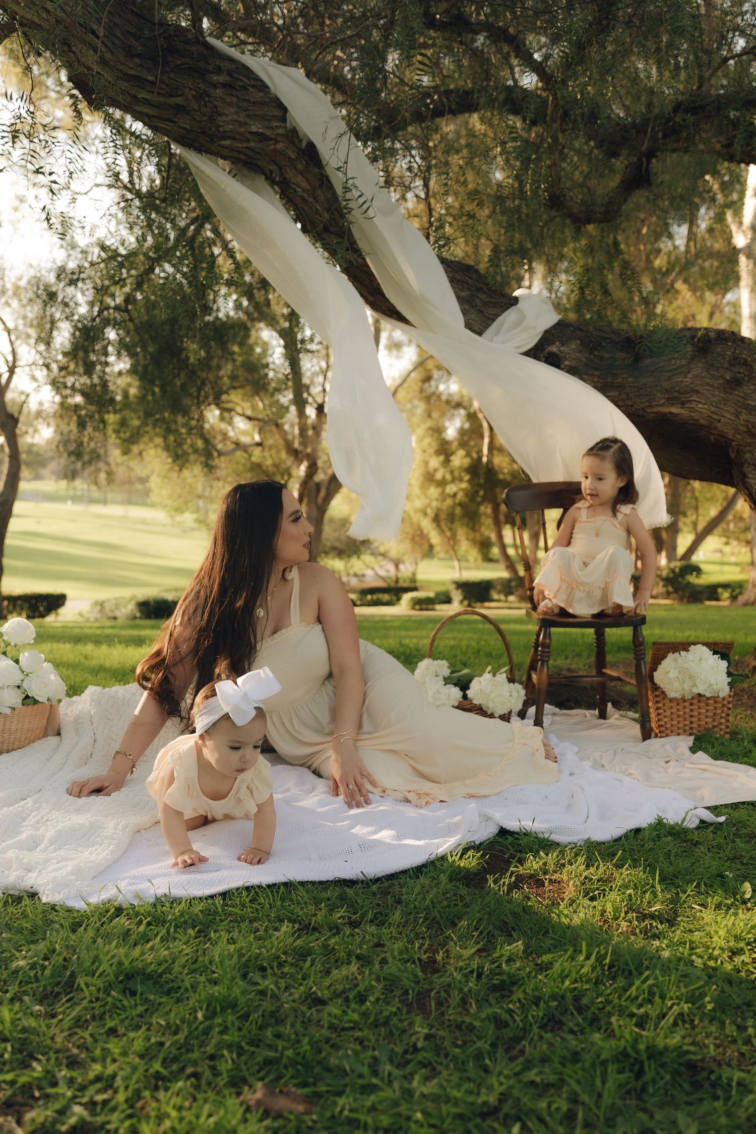 Mother in cream dress with two young children on white blanket under tree in park.
