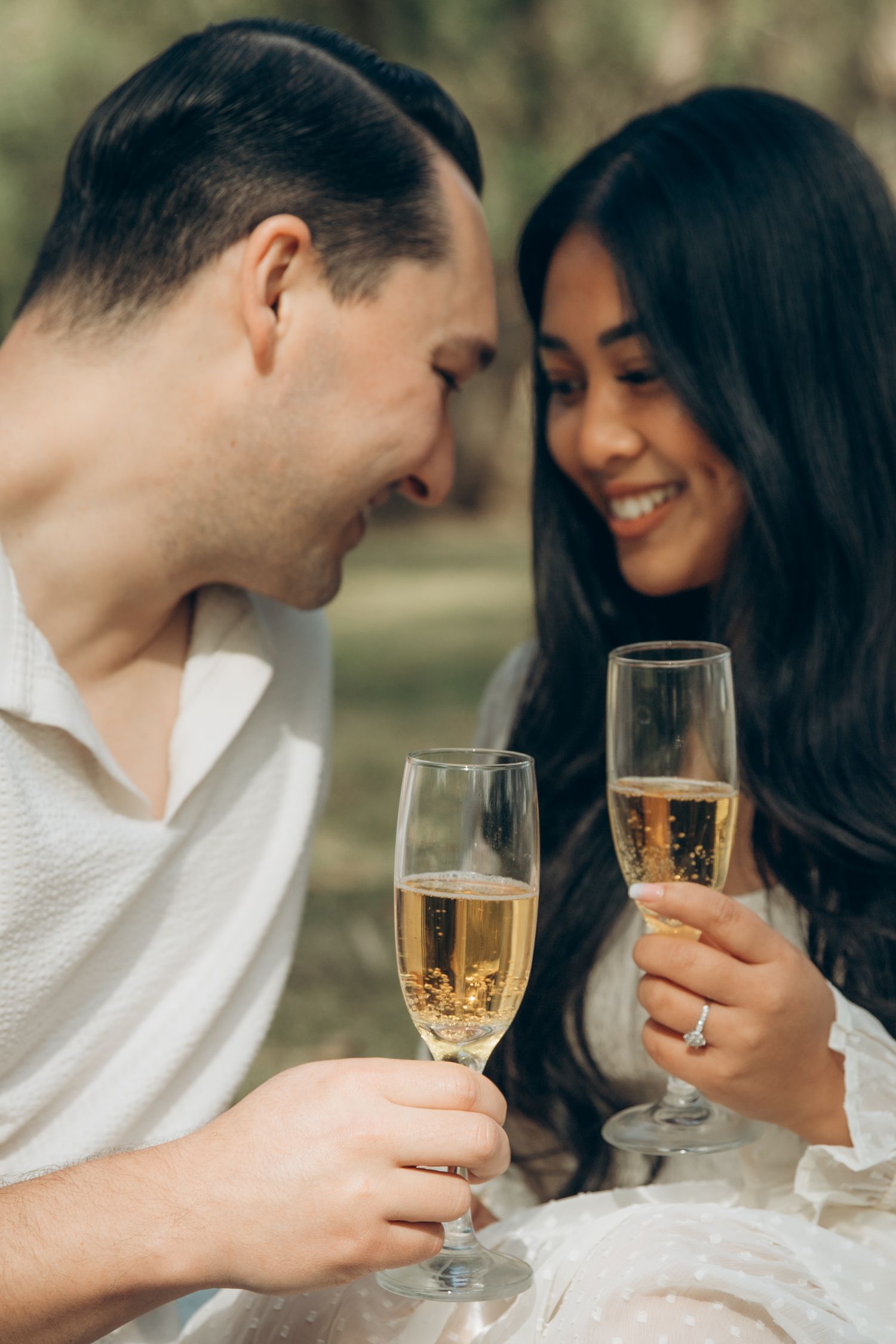 Smiling couple clinking champagne glasses together during an outdoor picnic, woman wearing an engagement ring.