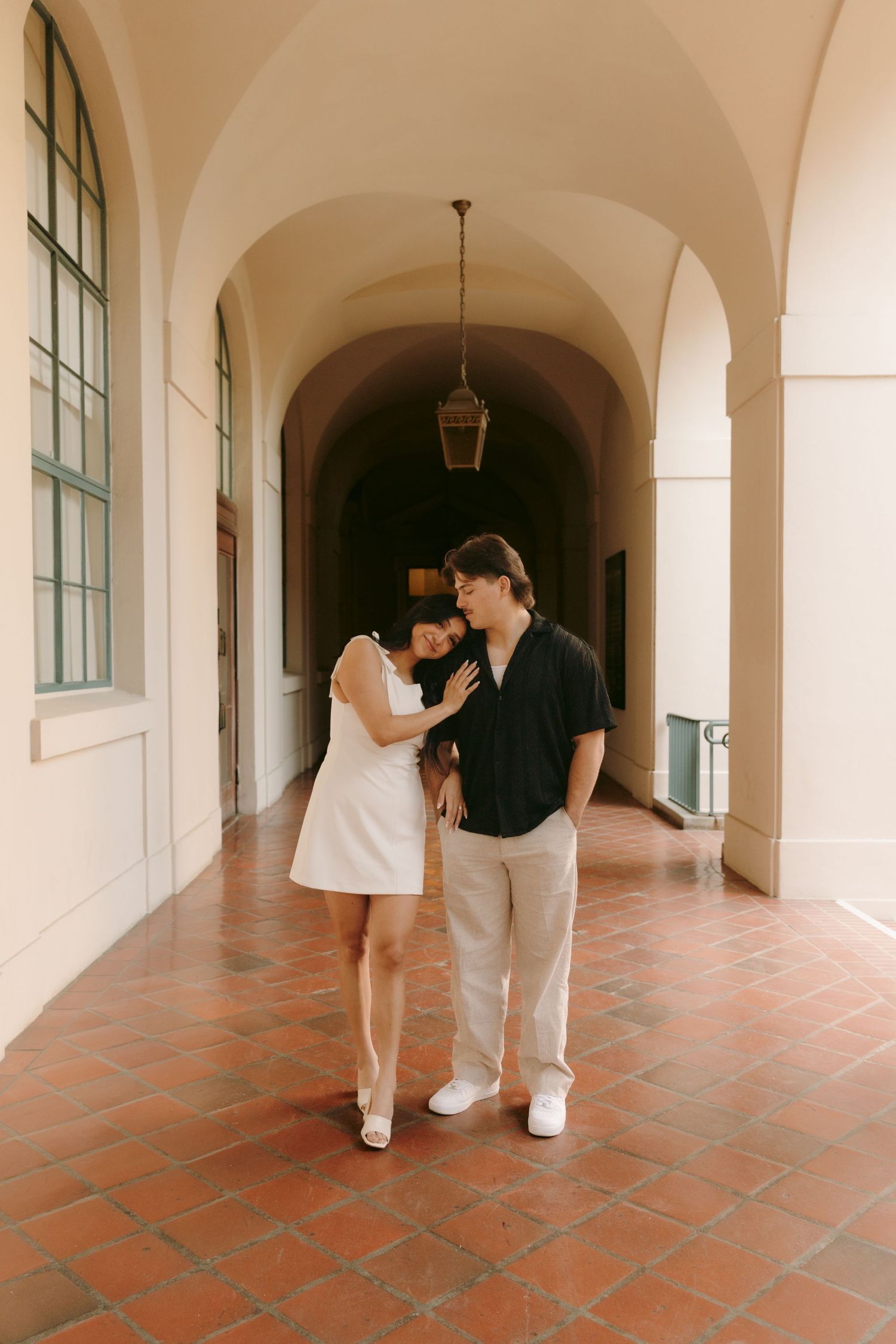 A couple shares a tender moment in a sunlit arched corridor with terracotta tile floors.