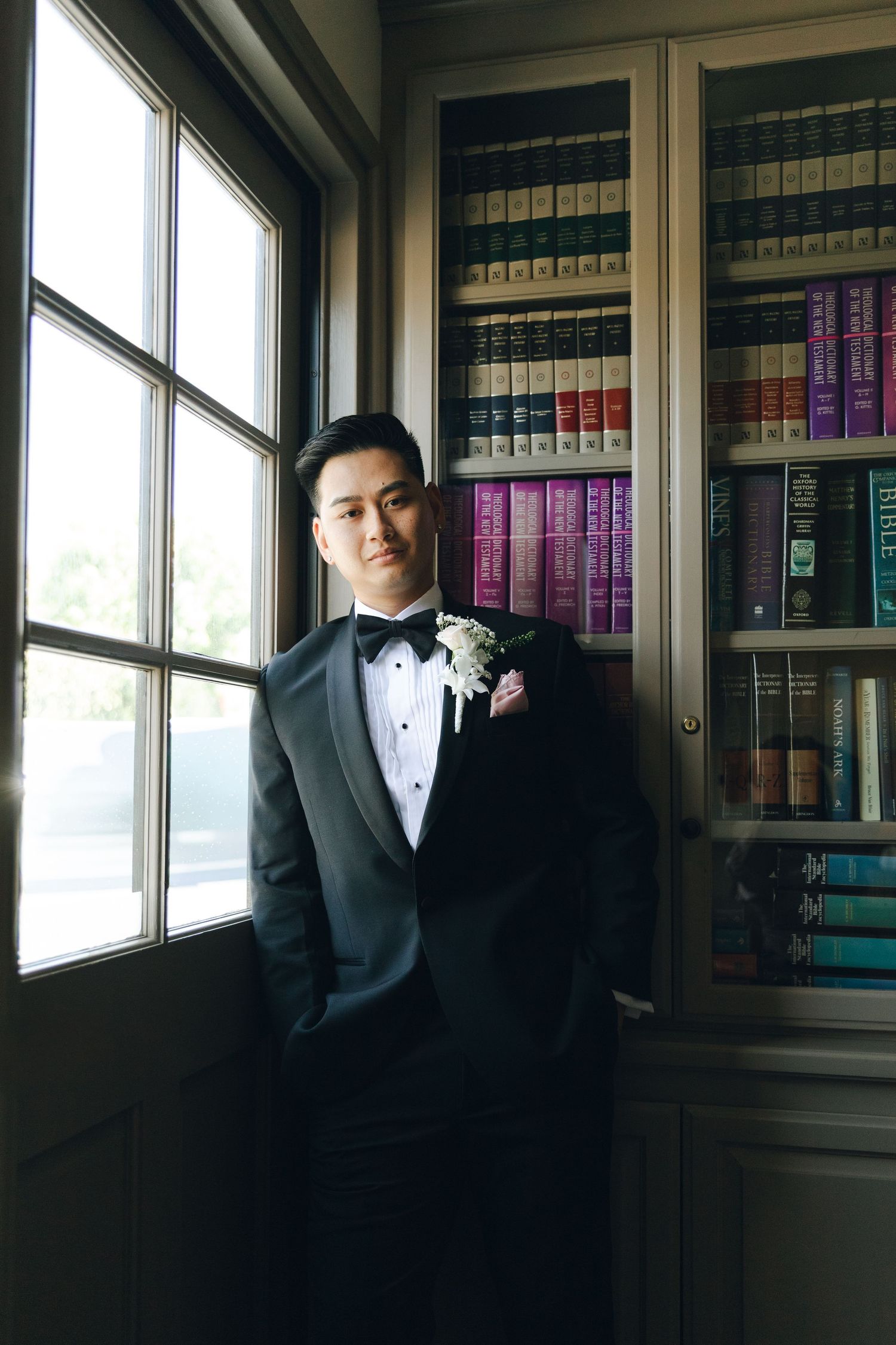 Dapper Asian groom in black tuxedo with boutonniere posing by bookshelf and window.