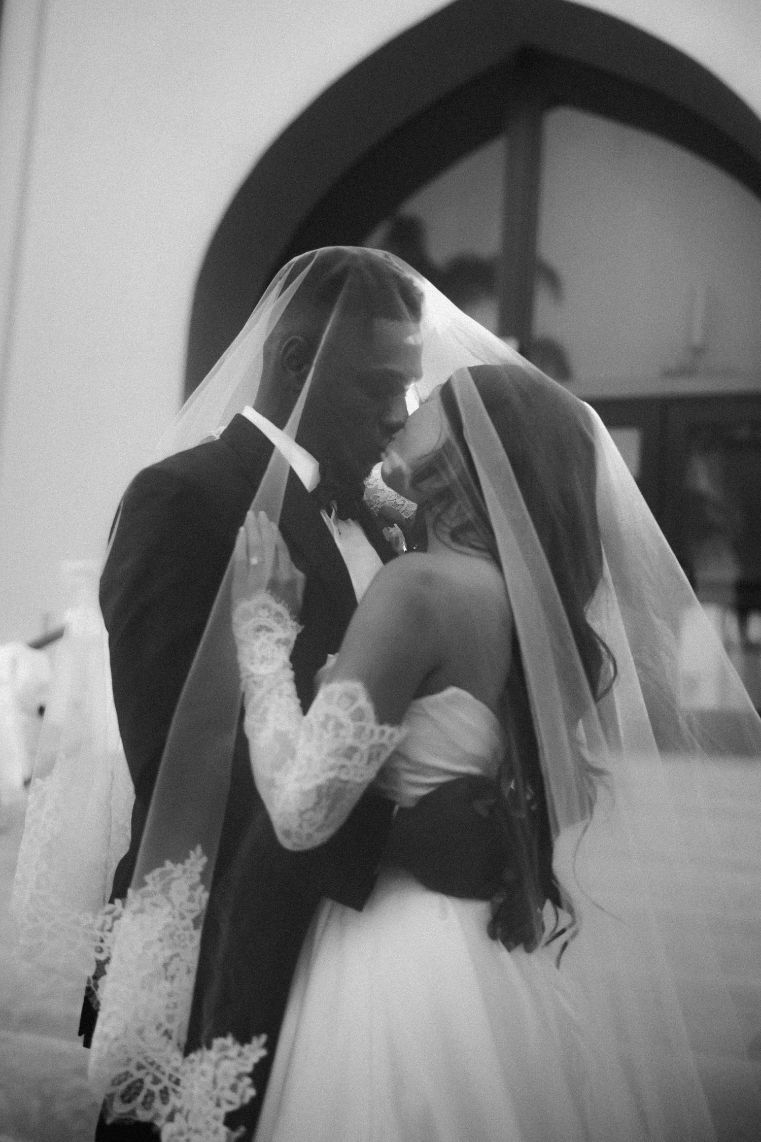 Black and white photo of bride and groom sharing a kiss under a lace wedding veil outside a church.