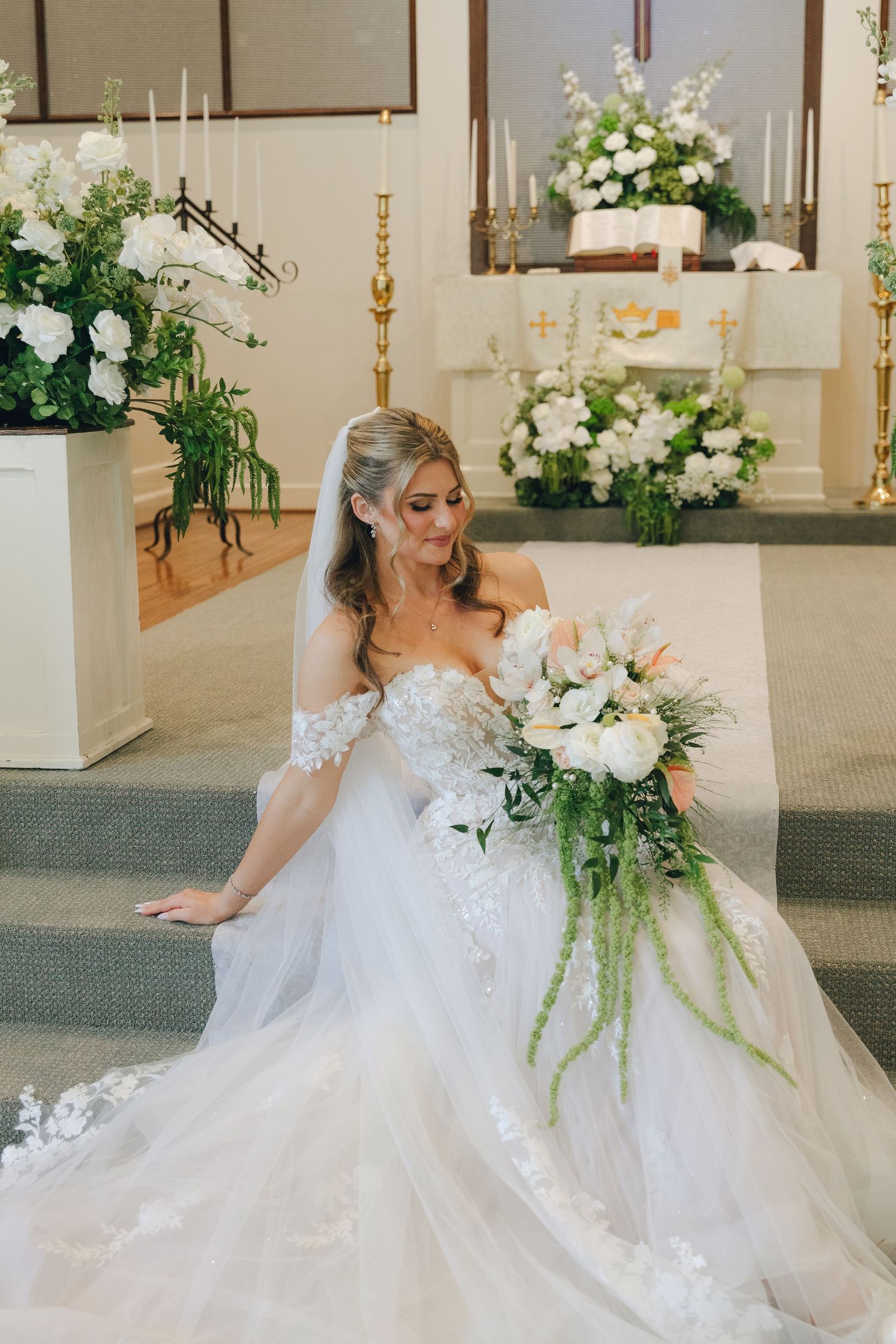 Bride in off-shoulder lace ball gown sits on church altar steps holding cascading white and green bouquet.