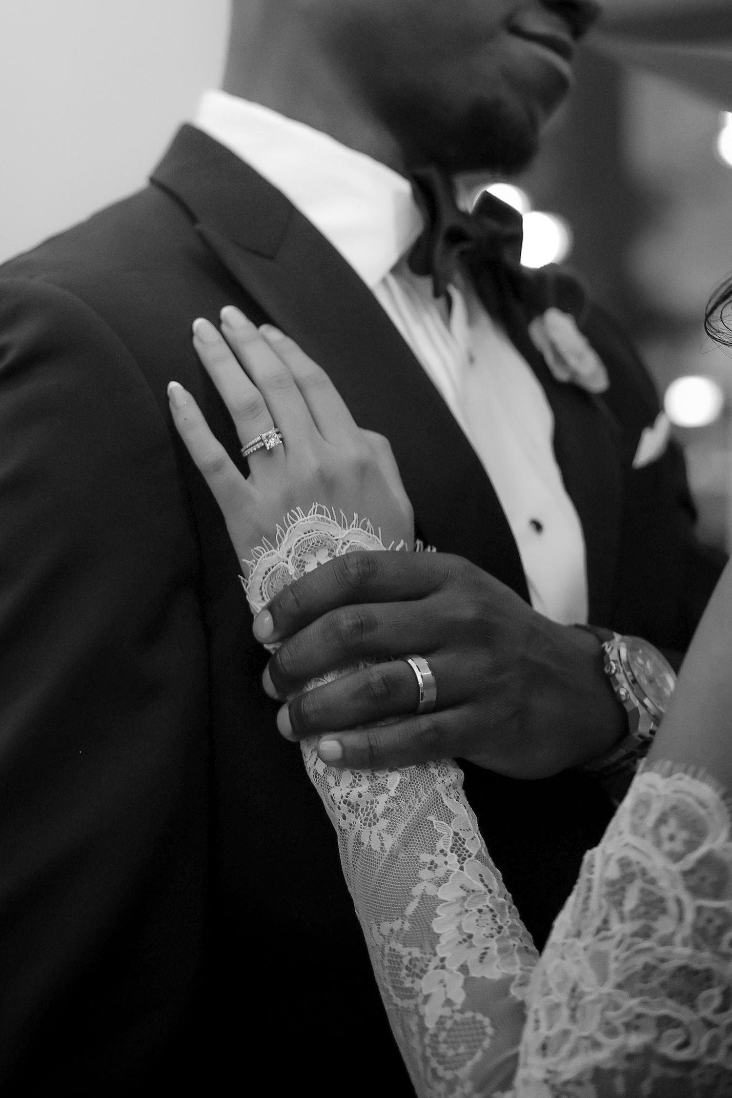Black and white photo of bride and groom holding hands showing wedding rings, bride in lace gown.