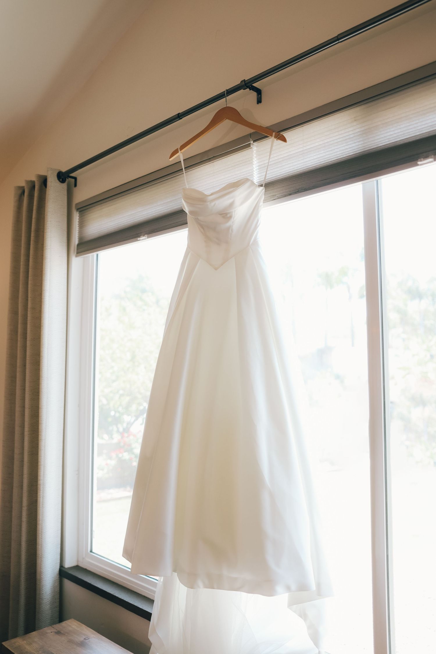 Elegant white strapless wedding gown hanging on wooden hanger in front of bright window.