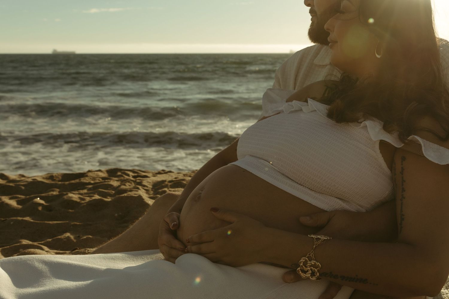 Close-up of pregnant woman's belly being lovingly held by partner on sunny beach.