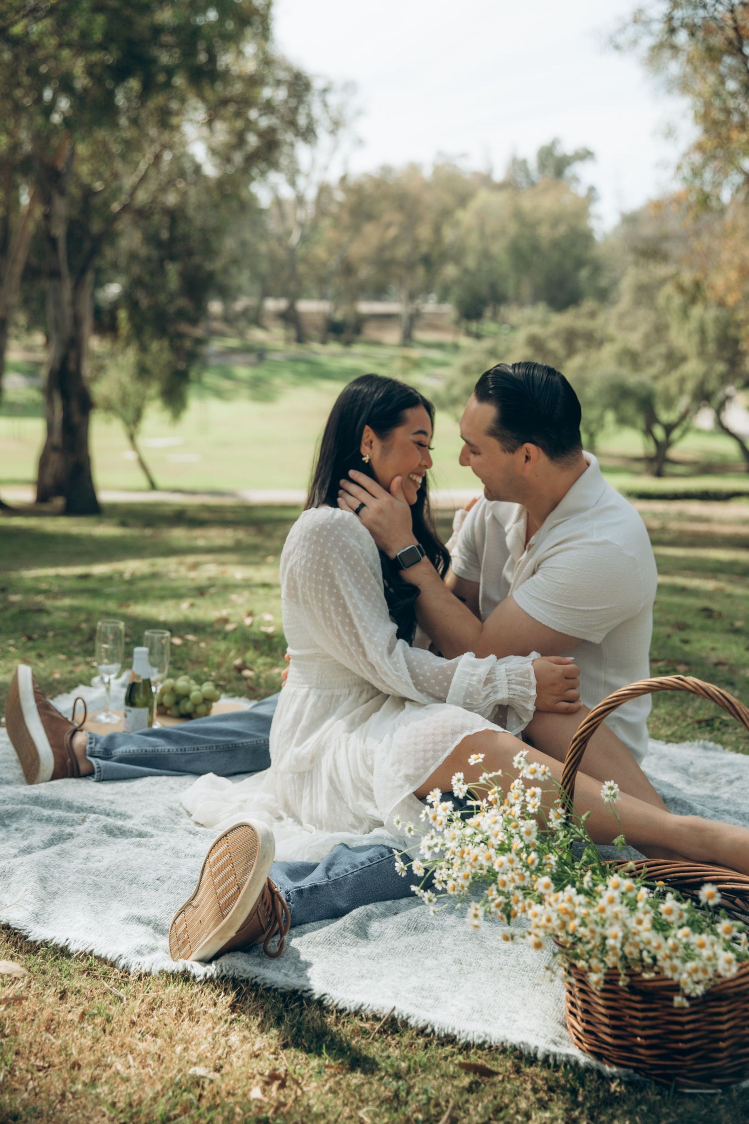 Romantic couple sharing a tender moment on a blanket during a sunny park picnic with daisies nearby.