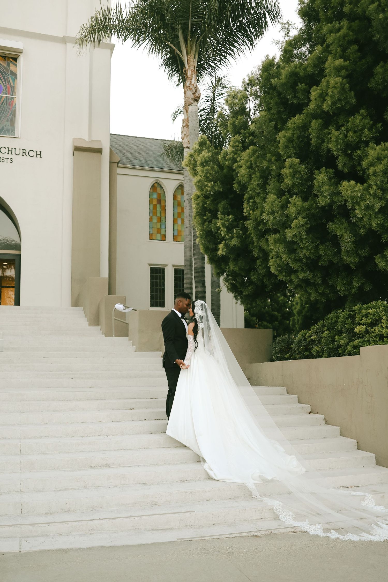 Bride and groom share a romantic moment on church steps, her long white veil trailing behind them.