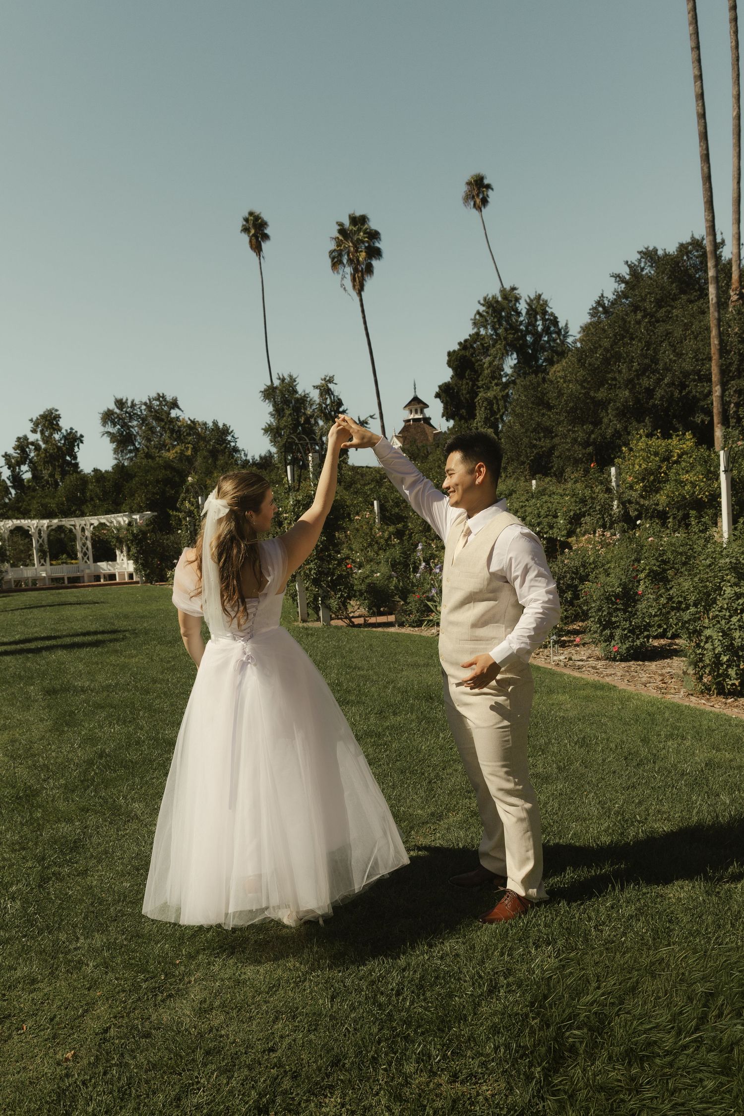 Bride and groom dancing joyfully on a green lawn with palm trees and a garden backdrop.