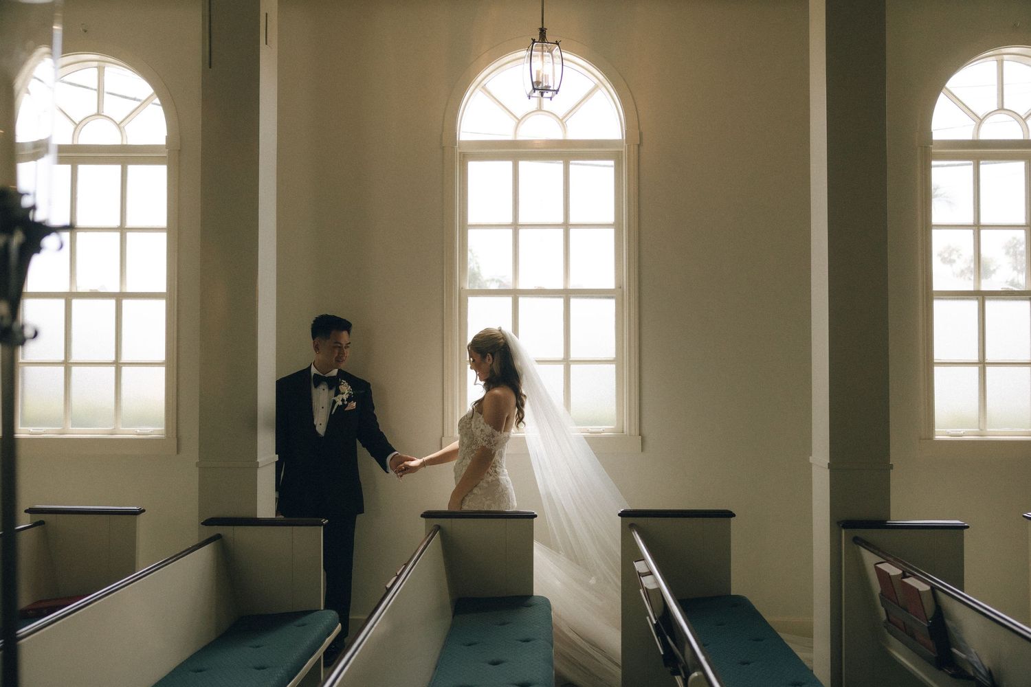 A bride and groom hold hands in a sunlit church with arched windows and blue carpet aisle on their wedding day.