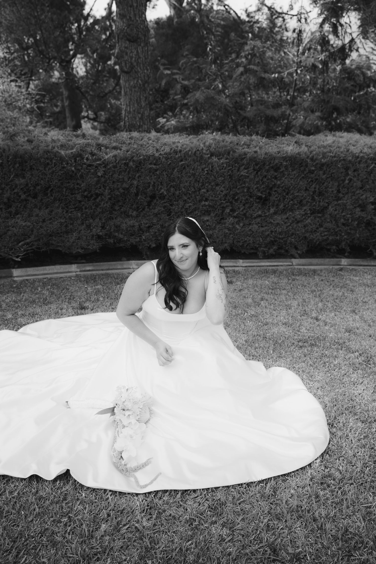 Bride in a flowing white ball gown sits gracefully on grass, holding bouquet, in a black and white outdoor portrait.