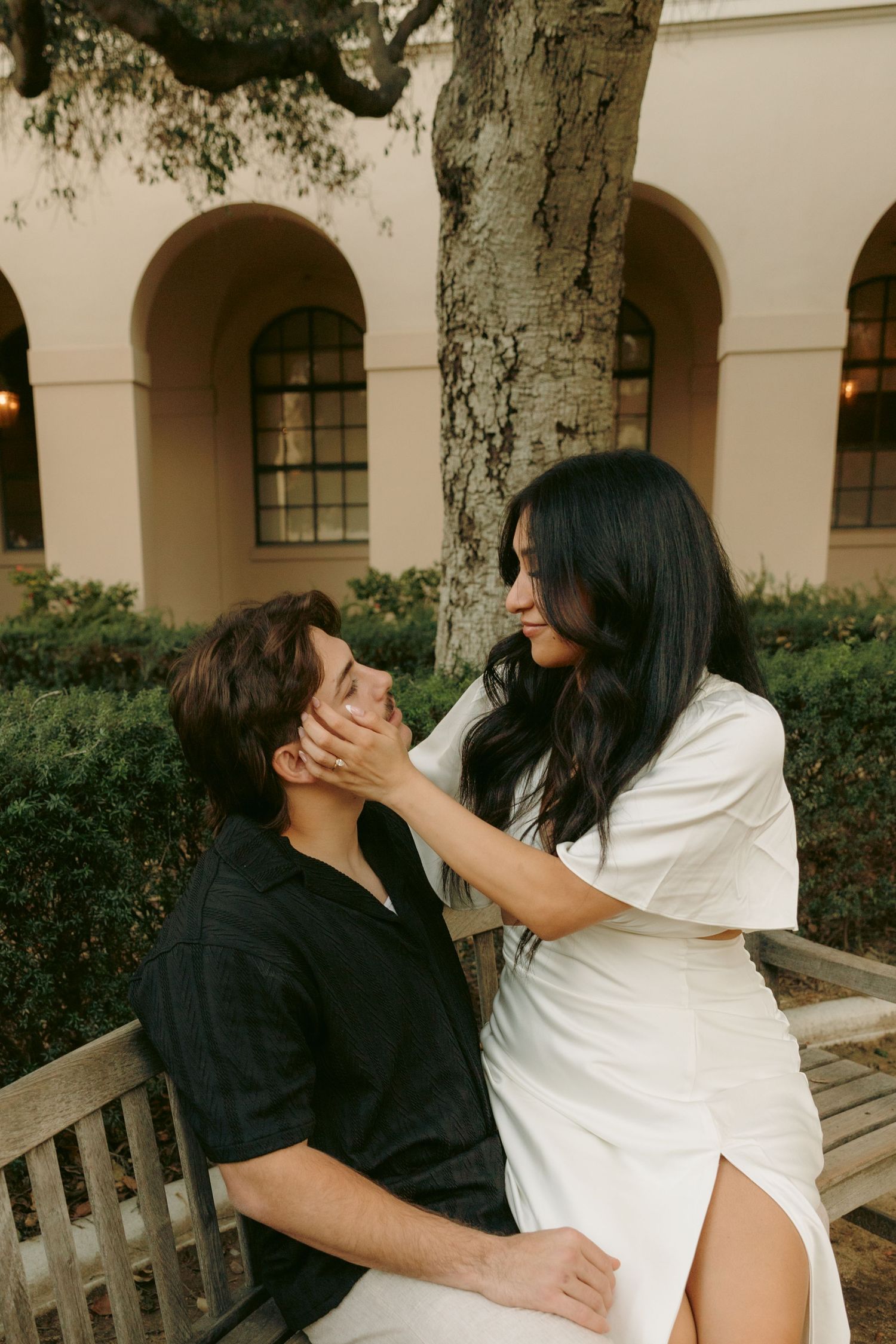 A woman tenderly holds a man's face while sitting together on a bench near arched building.