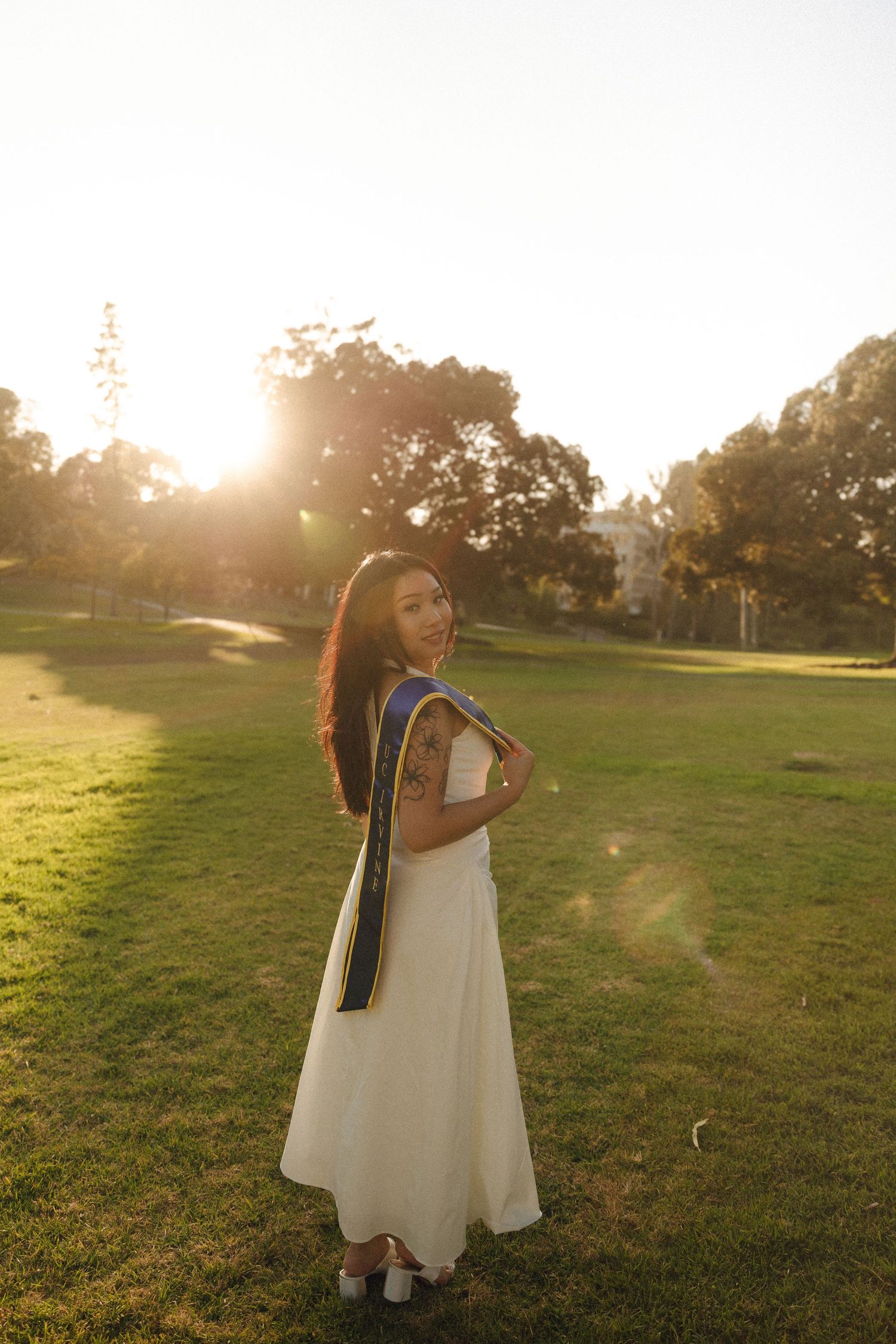 Young woman in white dress with blue graduation sash smiles over shoulder on sunny green lawn.