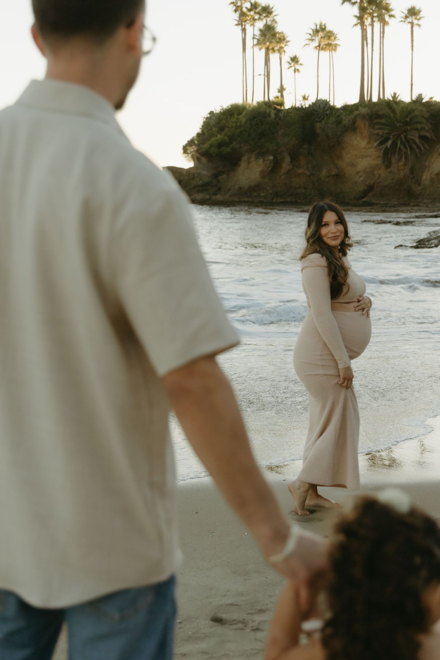 Pregnant woman in blush dress smiles on beach at sunset as man and child watch nearby.