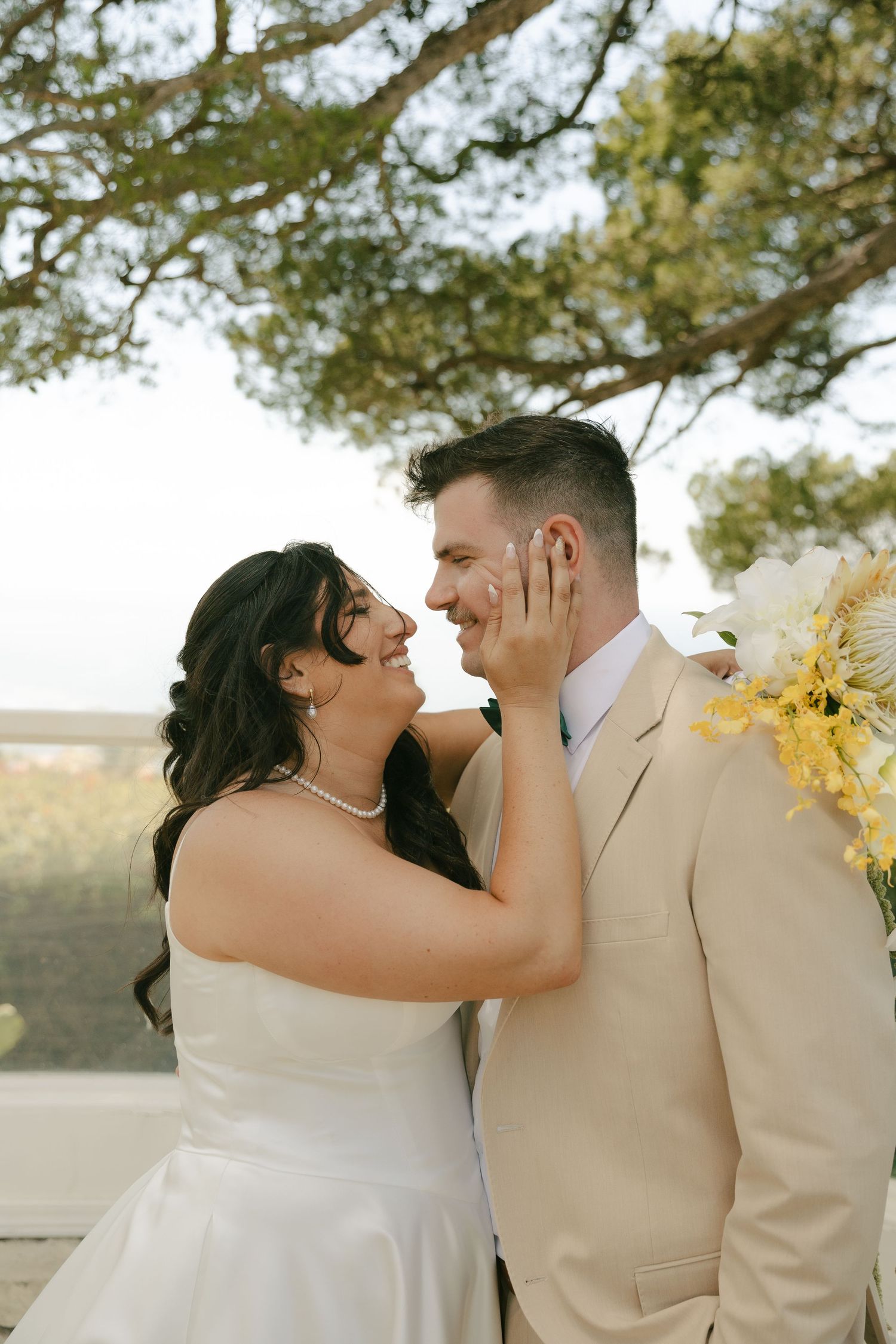 Bride and groom gazing at each other smiling under trees, groom in beige suit with yellow bouquet.
