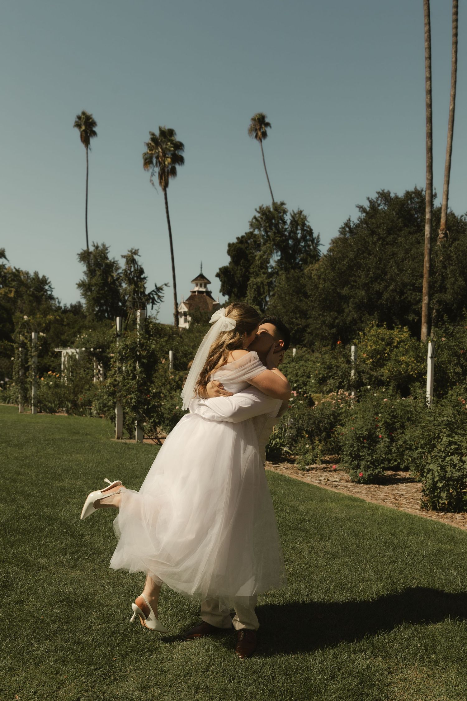 Groom lifting his bride in a white tulle wedding dress outdoors with tall palm trees in the background.