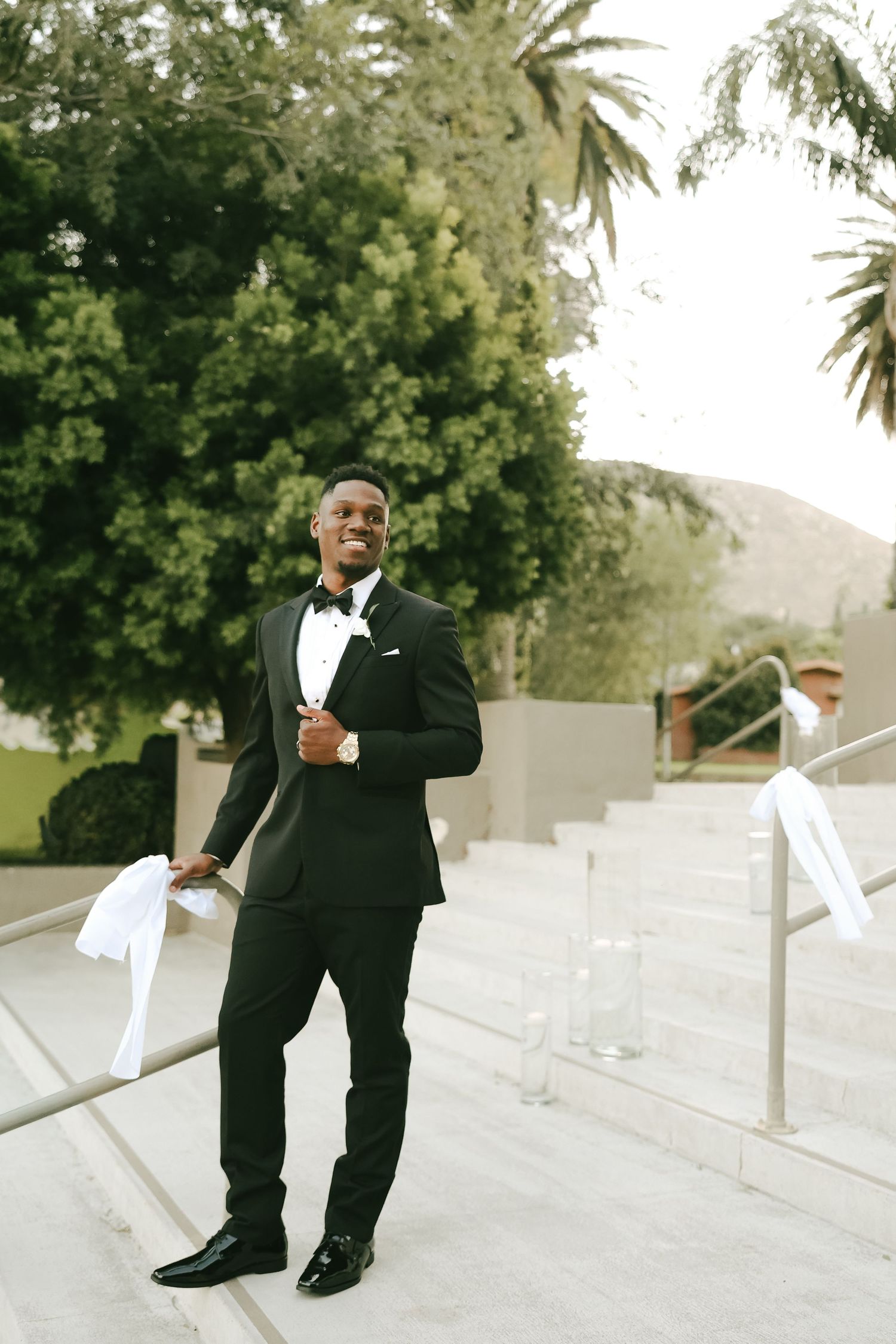 Handsome Black groom smiles confidently in classic black tuxedo with bow tie on outdoor venue steps with palm trees.