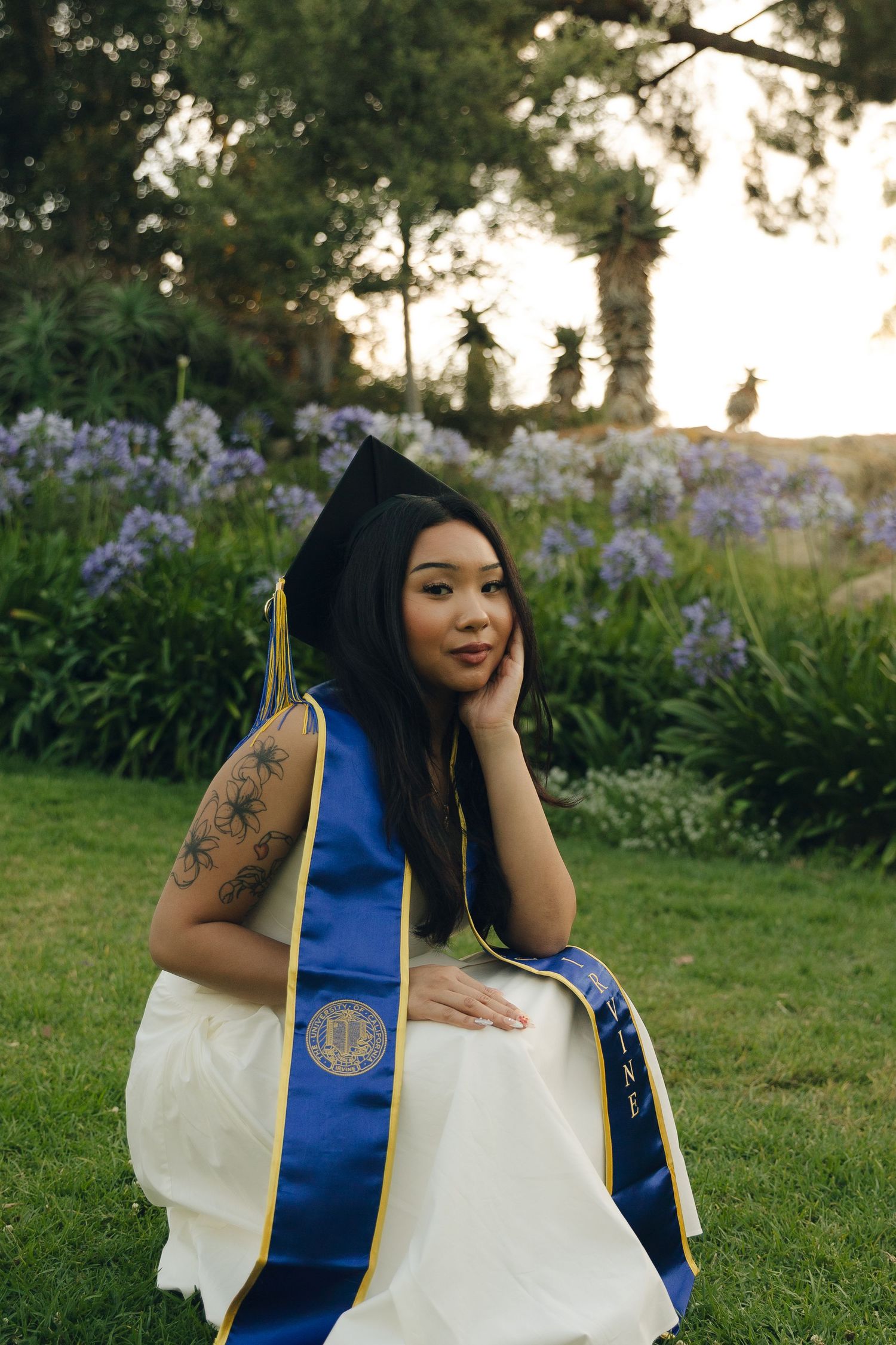 Female graduate in cap and blue sash sitting on grass surrounded by purple agapanthus at sunset.