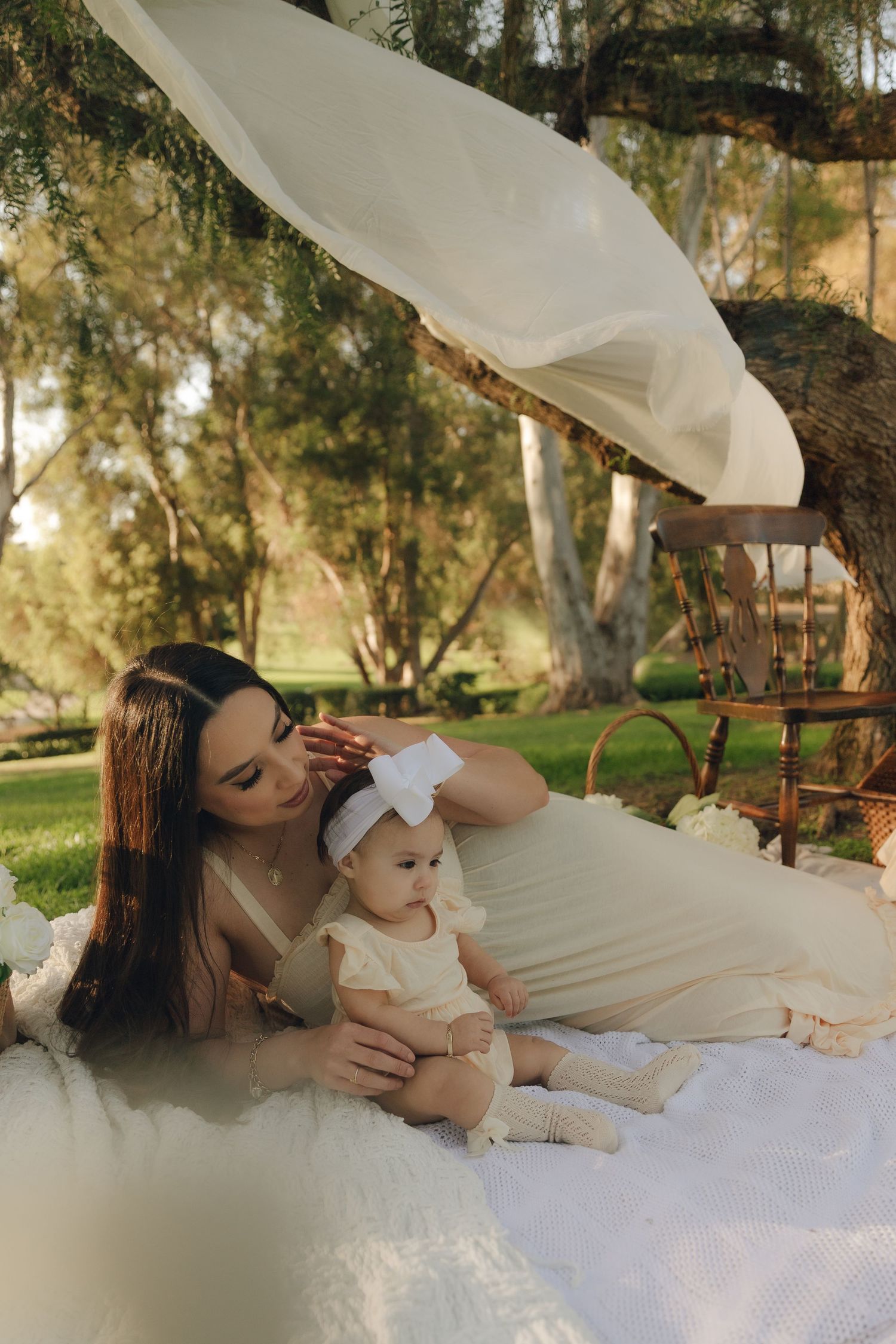 Mother in cream dress tenderly adjusting baby's white bow headband on outdoor picnic blanket.