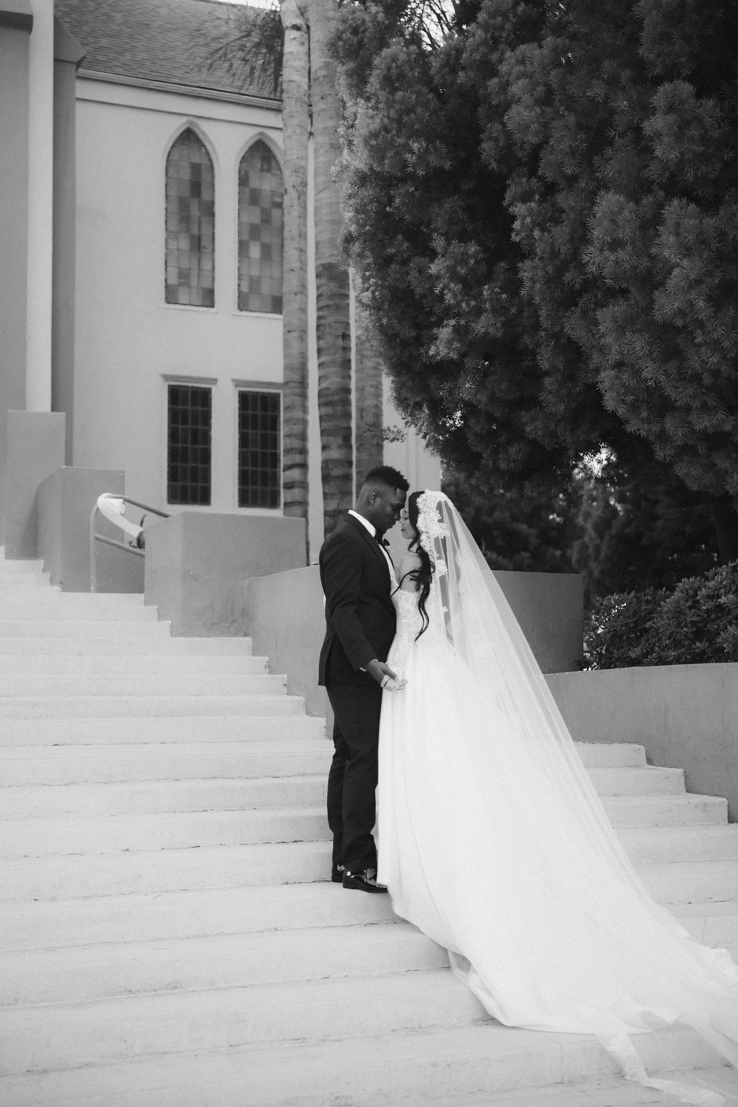 Black and white portrait of bride and groom sharing an intimate moment on church steps with long flowing veil.