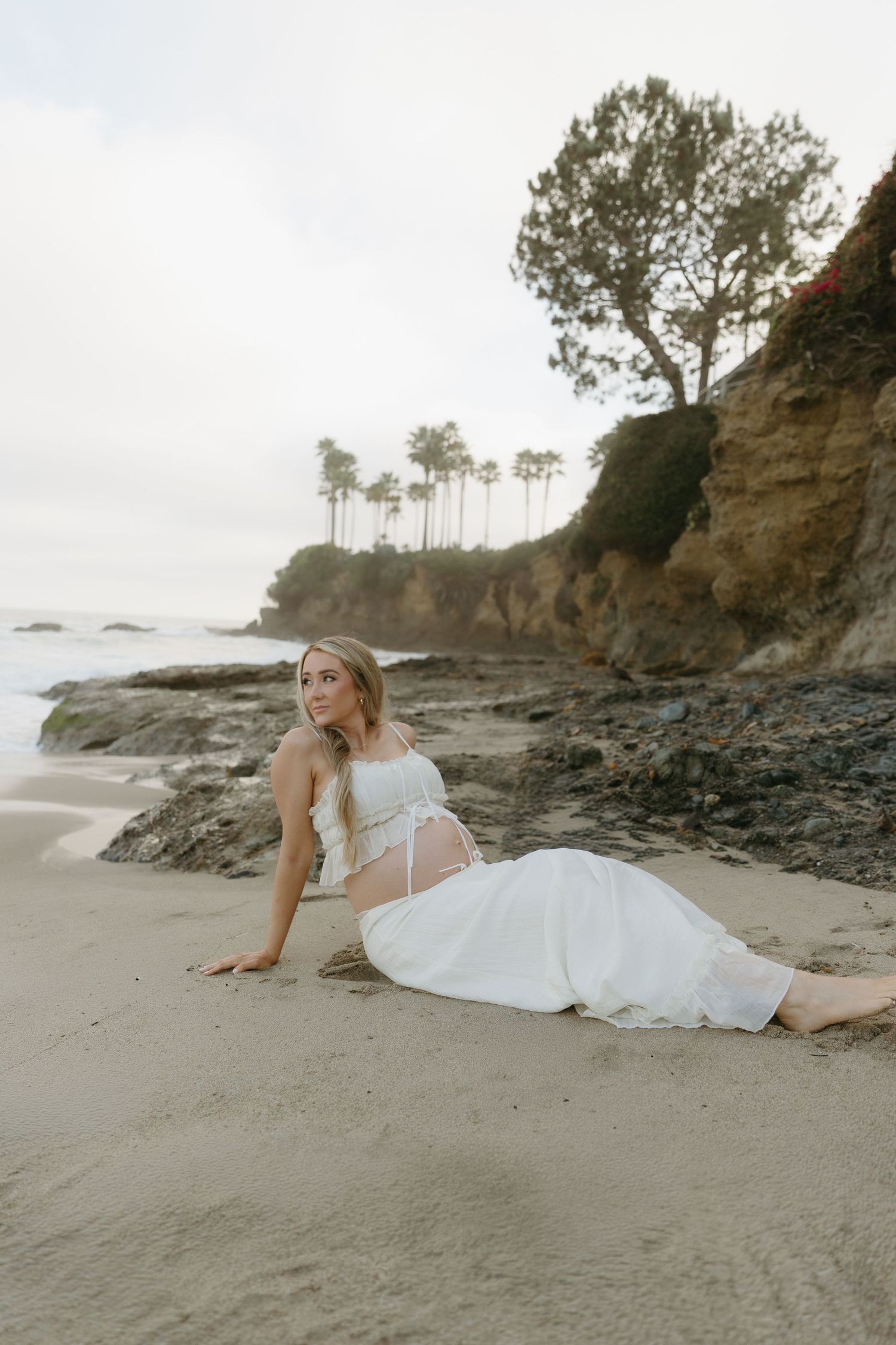 Pregnant blonde woman in white dress sitting gracefully on sandy beach with rocky cliffs and palm trees behind.